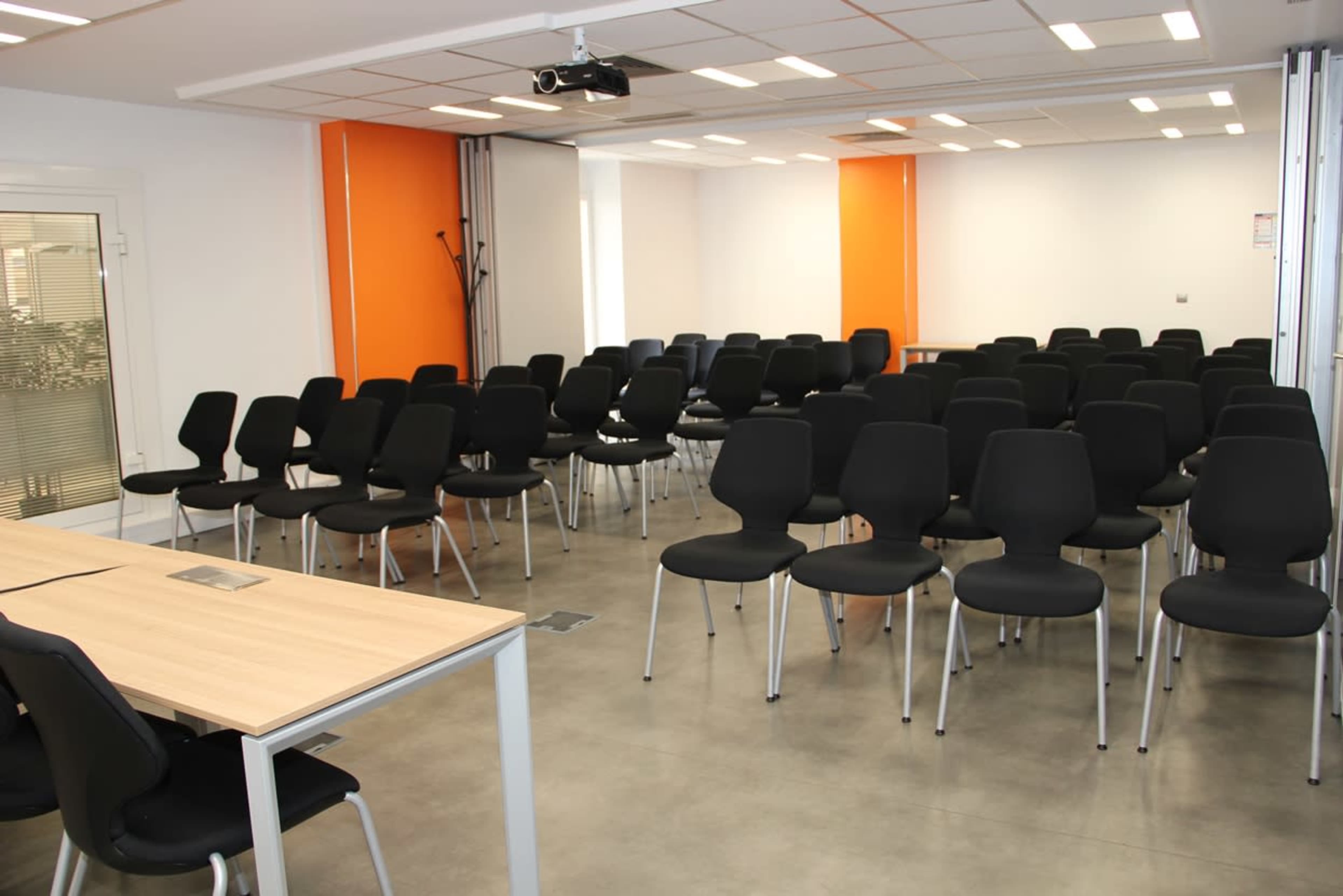 A conference room set up with rows of black chairs facing a desk and projector.