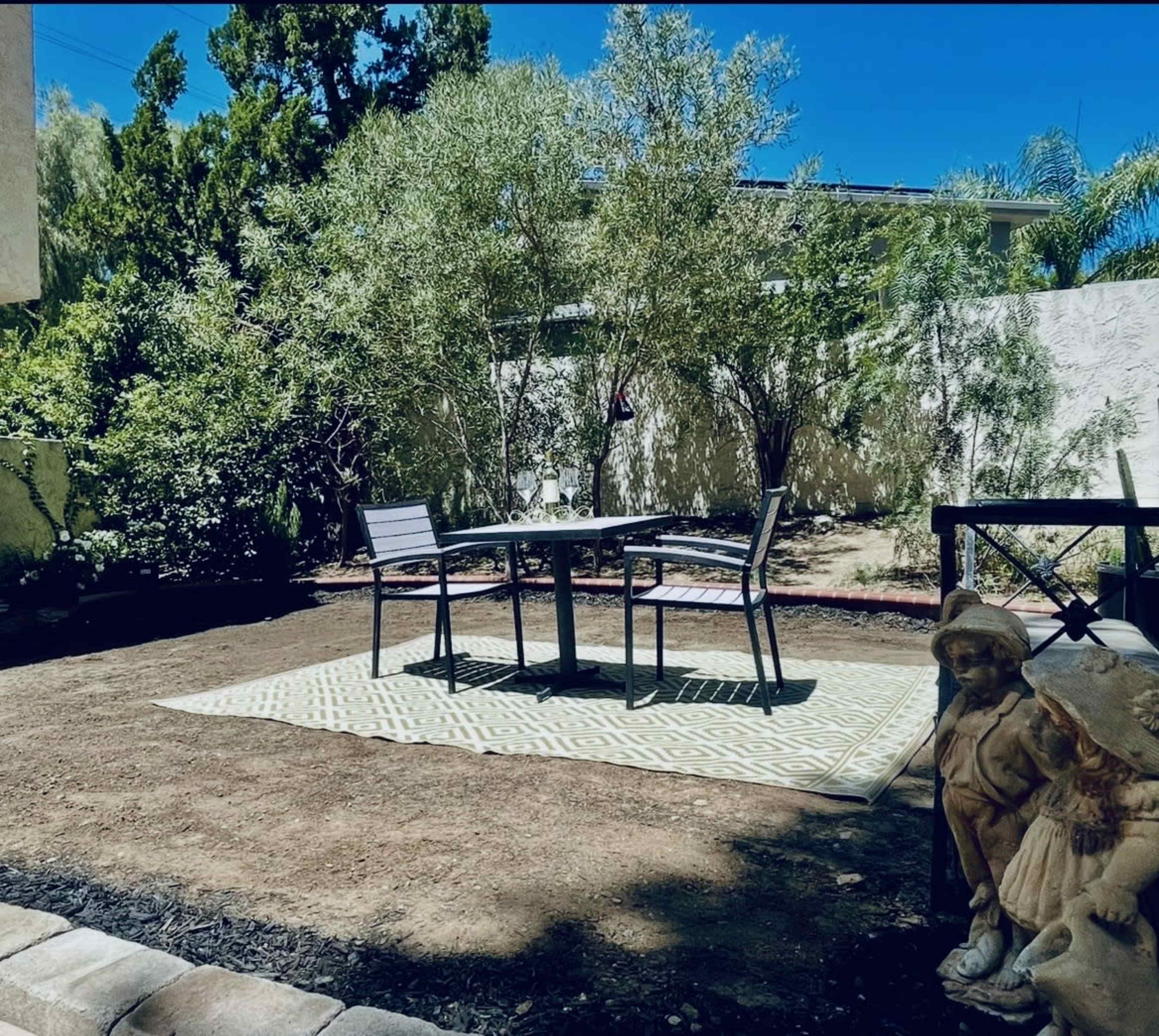The image shows a secluded outdoor patio area with a table and chairs set on a patterned rug, surrounded by greenery and a statue.