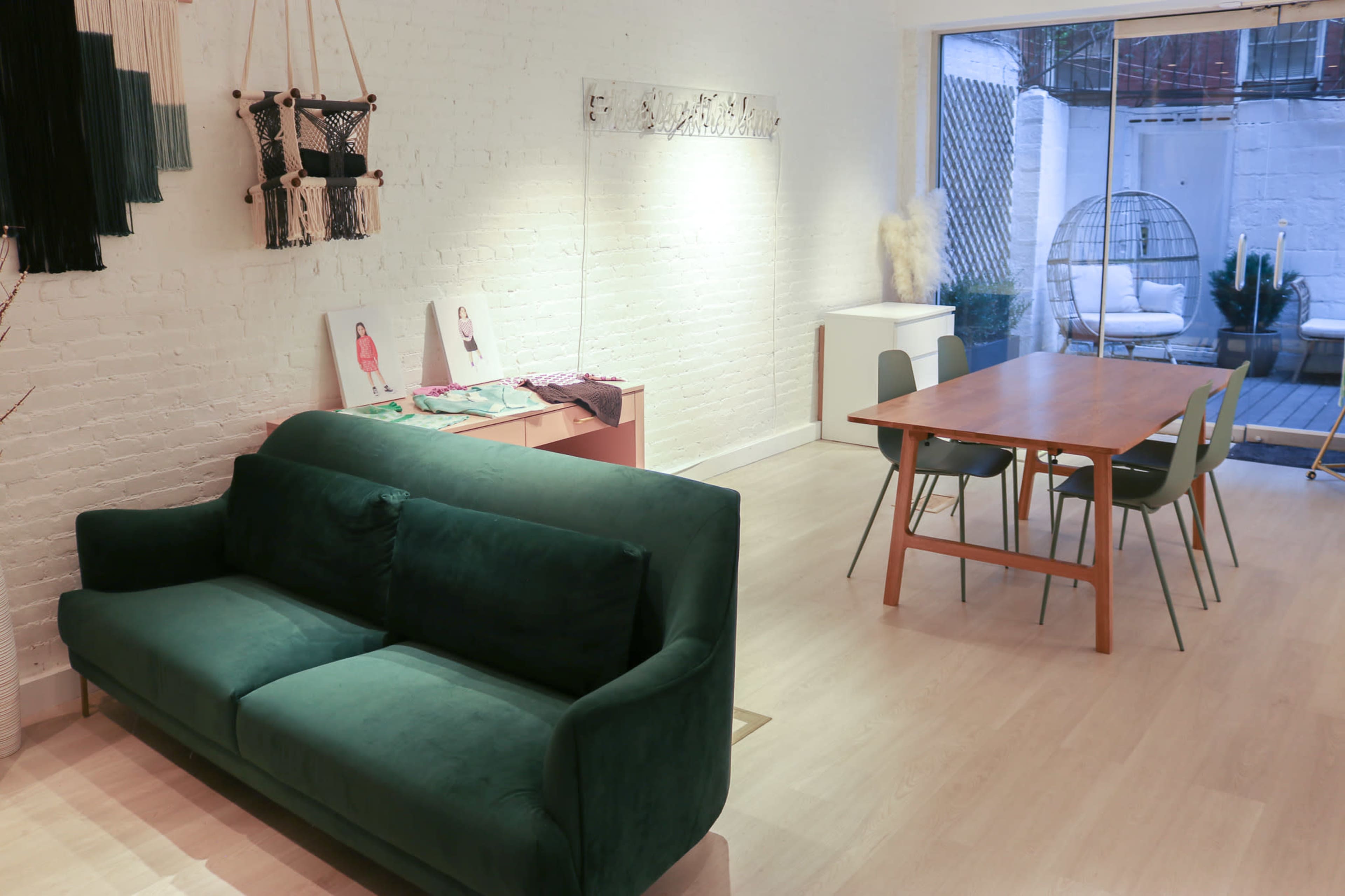 A bright interior featuring a green sofa, a wooden dining table with chairs, and a small outdoor area visible through a large window.