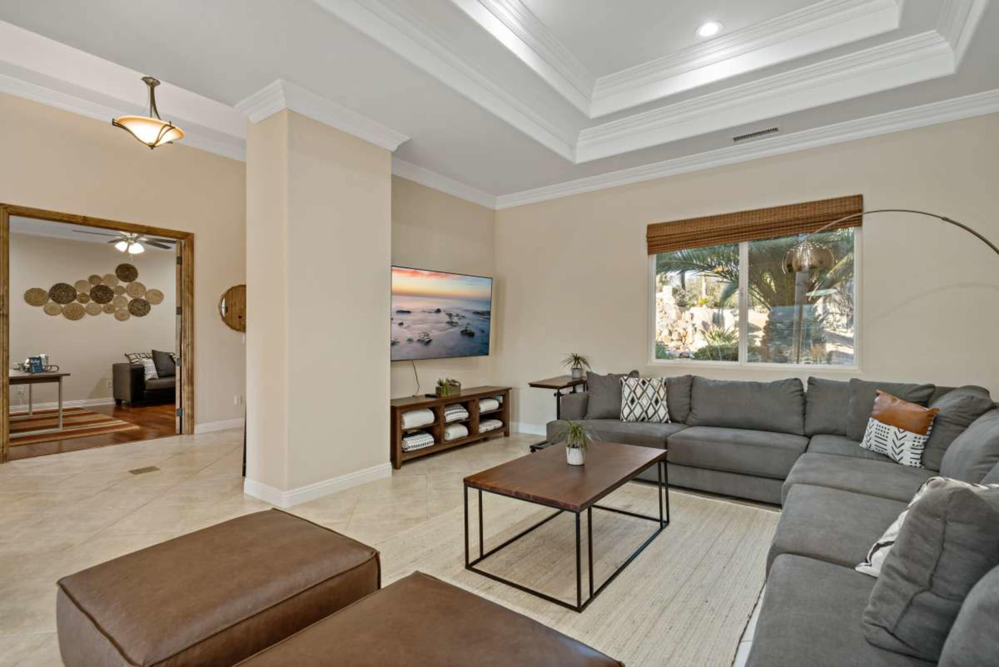 The image shows a spacious living room with a large sectional sofa, a coffee table, and a mounted TV on the wall, alongside a large window with views of outdoor greenery.