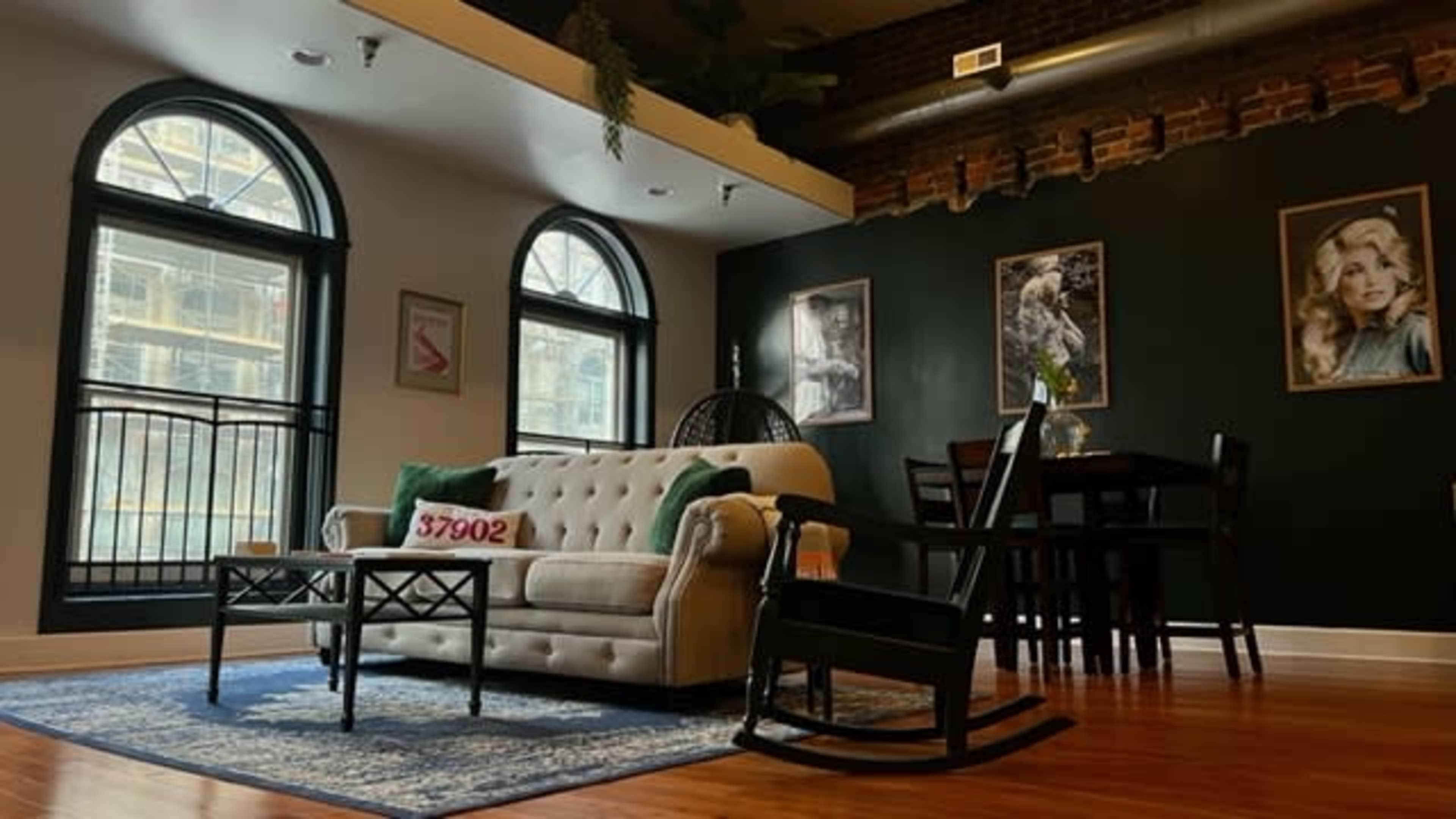 A living room with a white tufted sofa, a rocking chair, and a dining table, framed by large arched windows and brick walls.