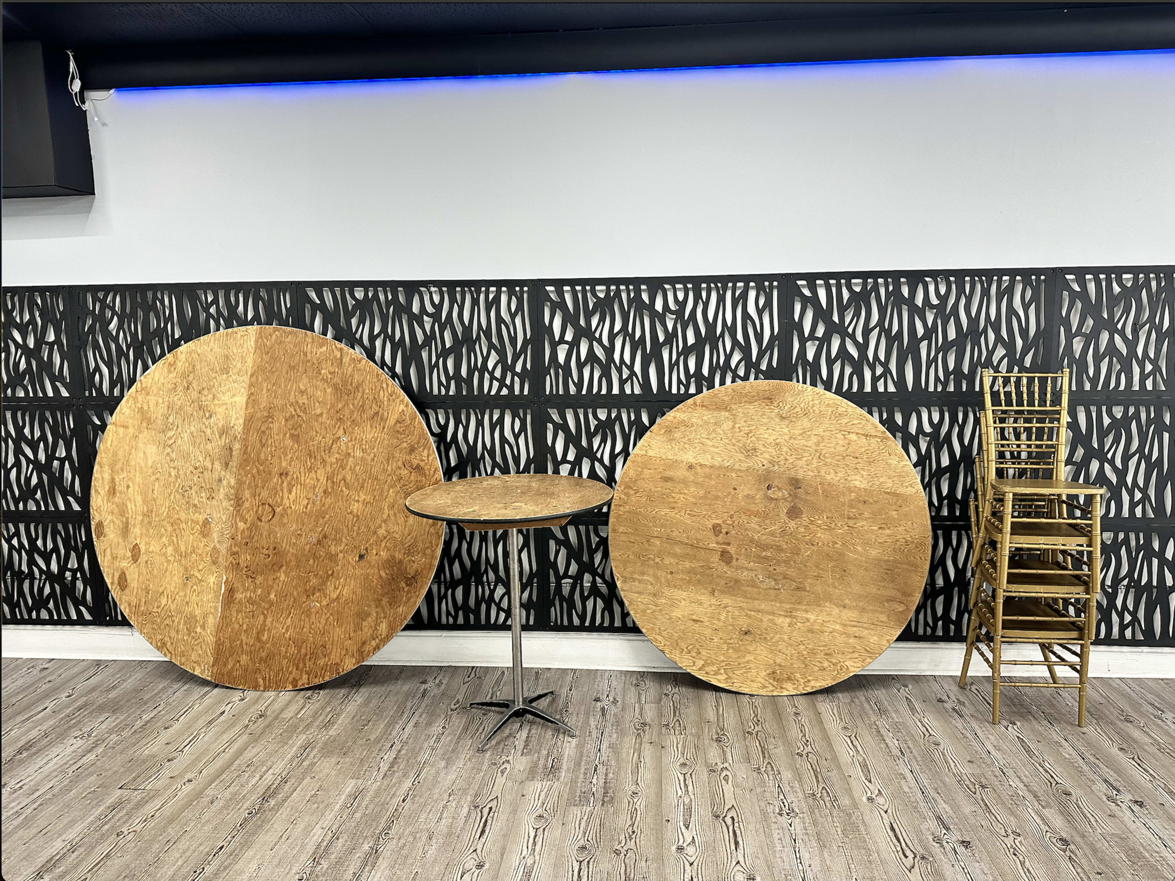 A circular wooden table is positioned between two large round wooden tops against a patterned wall, with a stack of gold-colored chairs nearby.