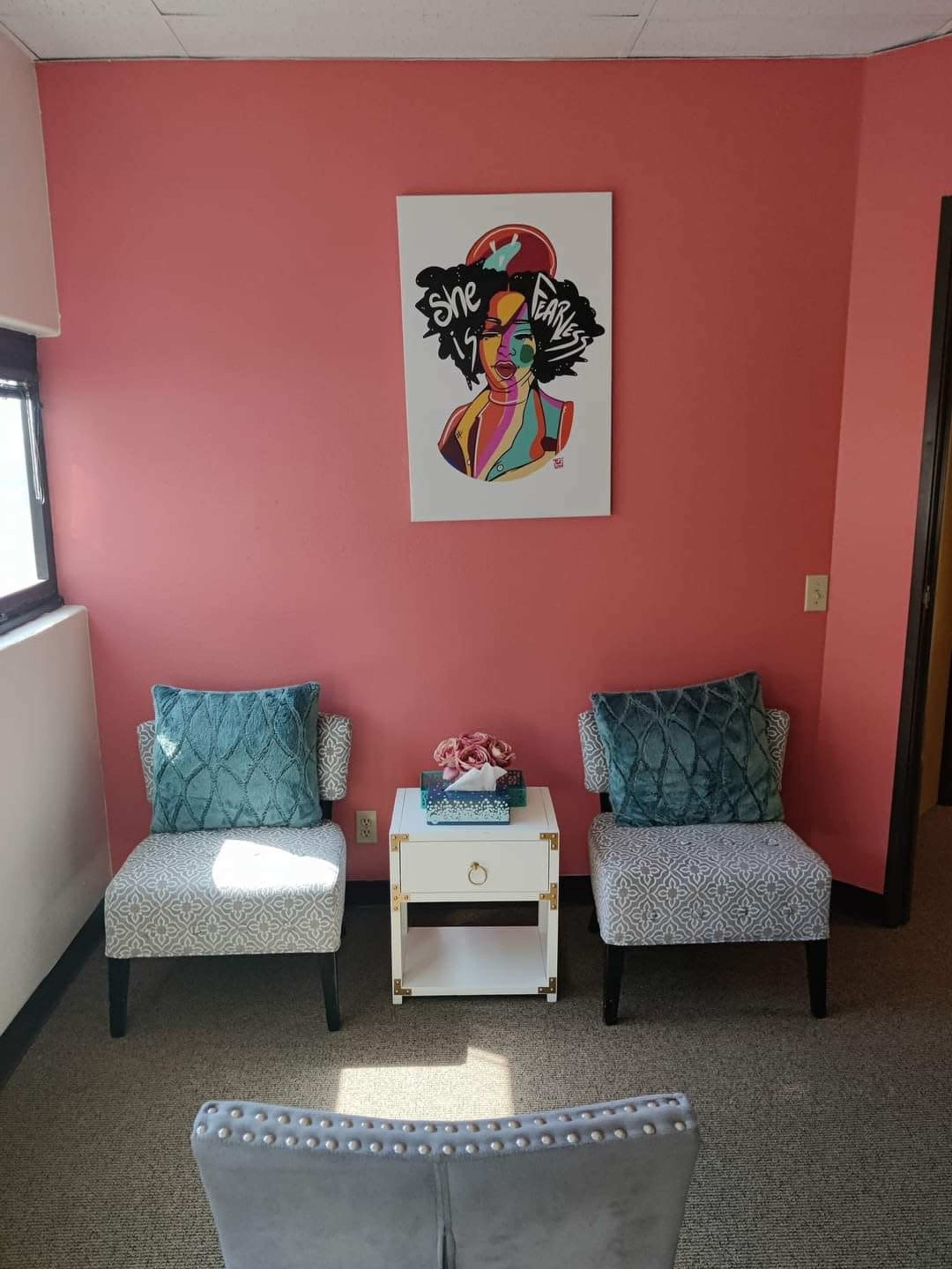 A small, brightly colored room with two patterned chairs facing a white table, adorned with a floral centerpiece and a colorful artwork on the pink wall.