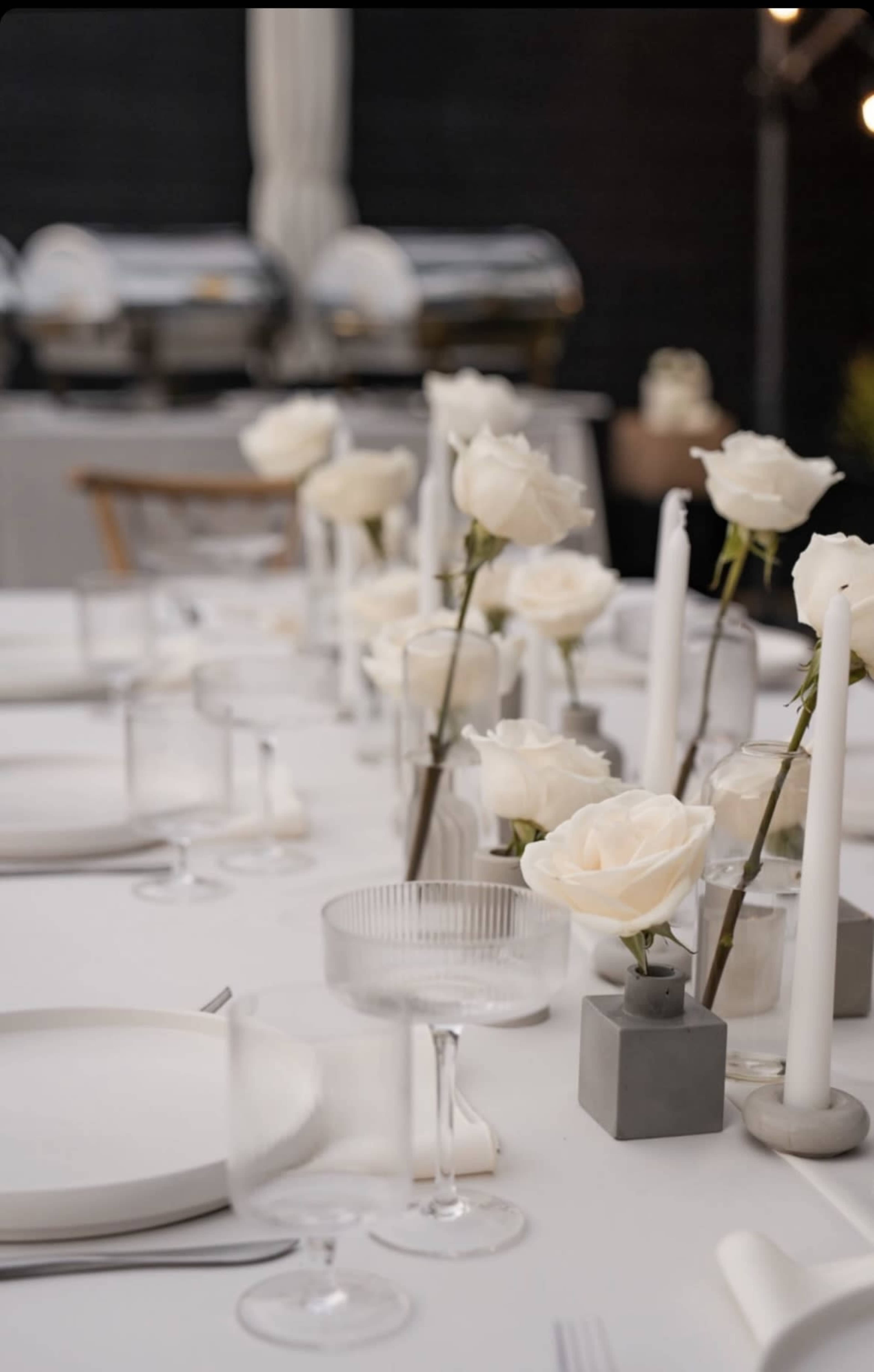A long dining table is set with white dishes, clear glasses, and centerpieces of white roses alongside white candles in minimalist holders.