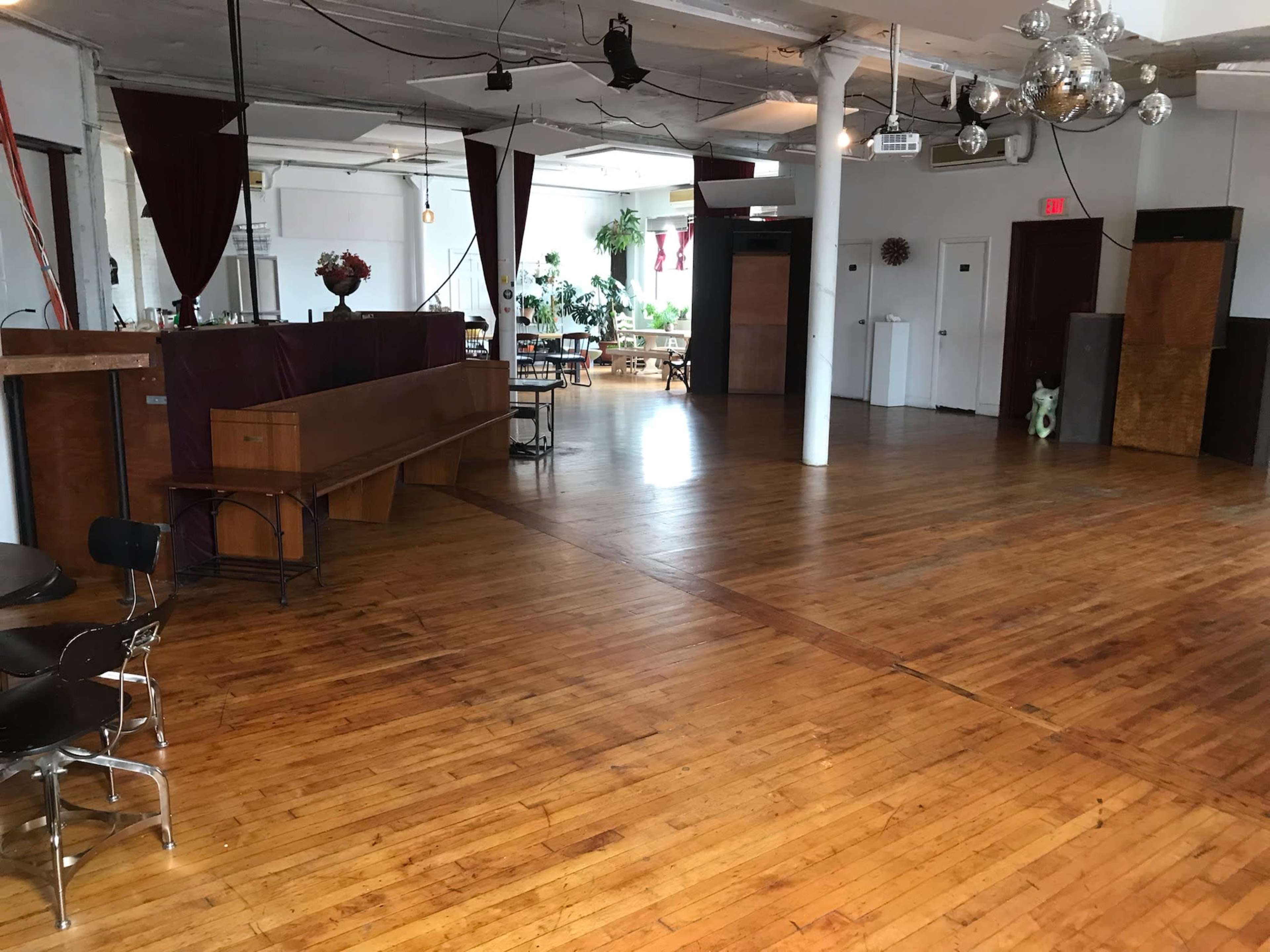 The interior space features polished wooden floors, a bar area, tables and chairs, with large windows and greenery in the background.