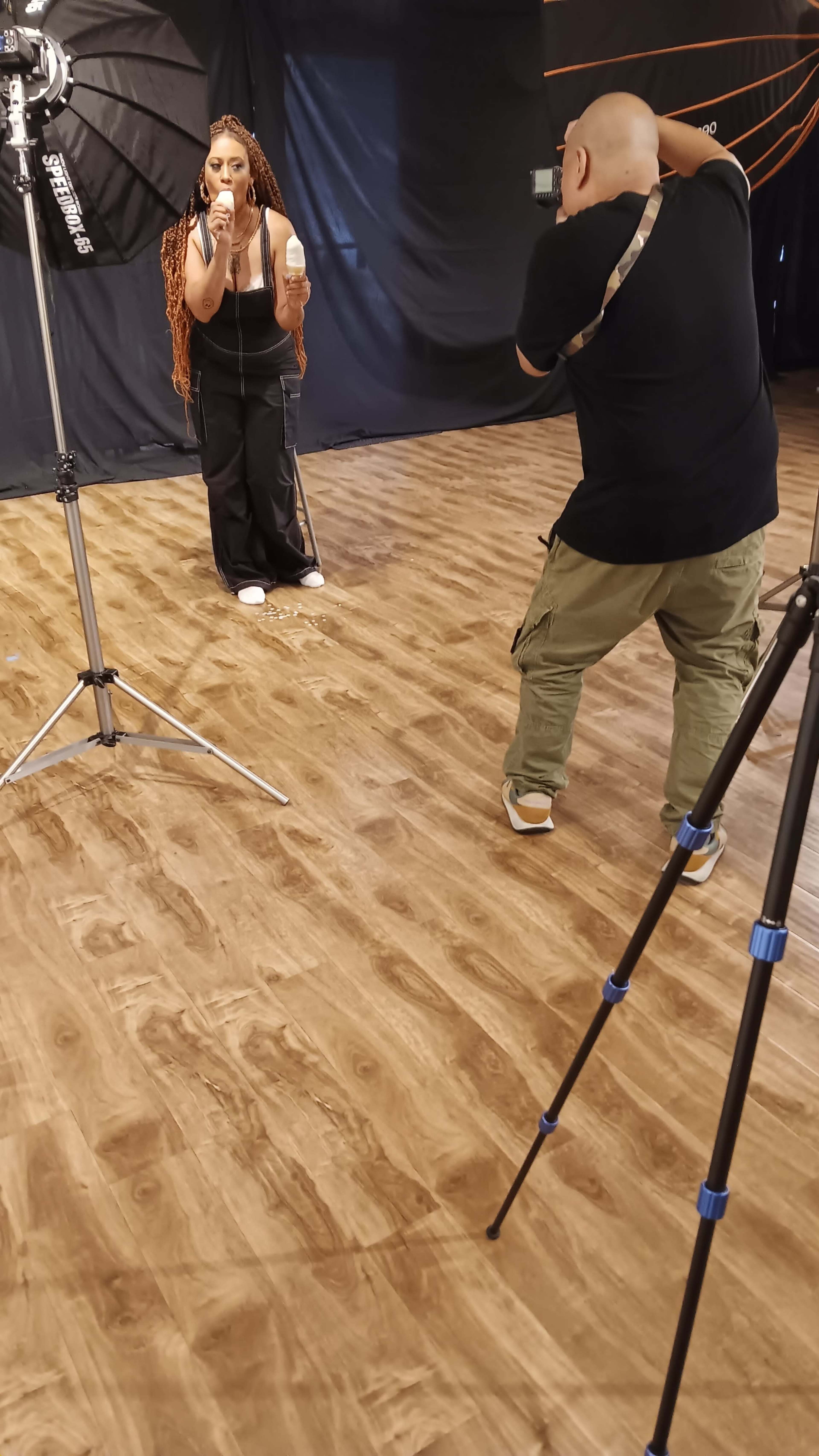 A photographer is capturing pictures of a woman with long hair in a studio setting, surrounded by lighting equipment and a black backdrop.