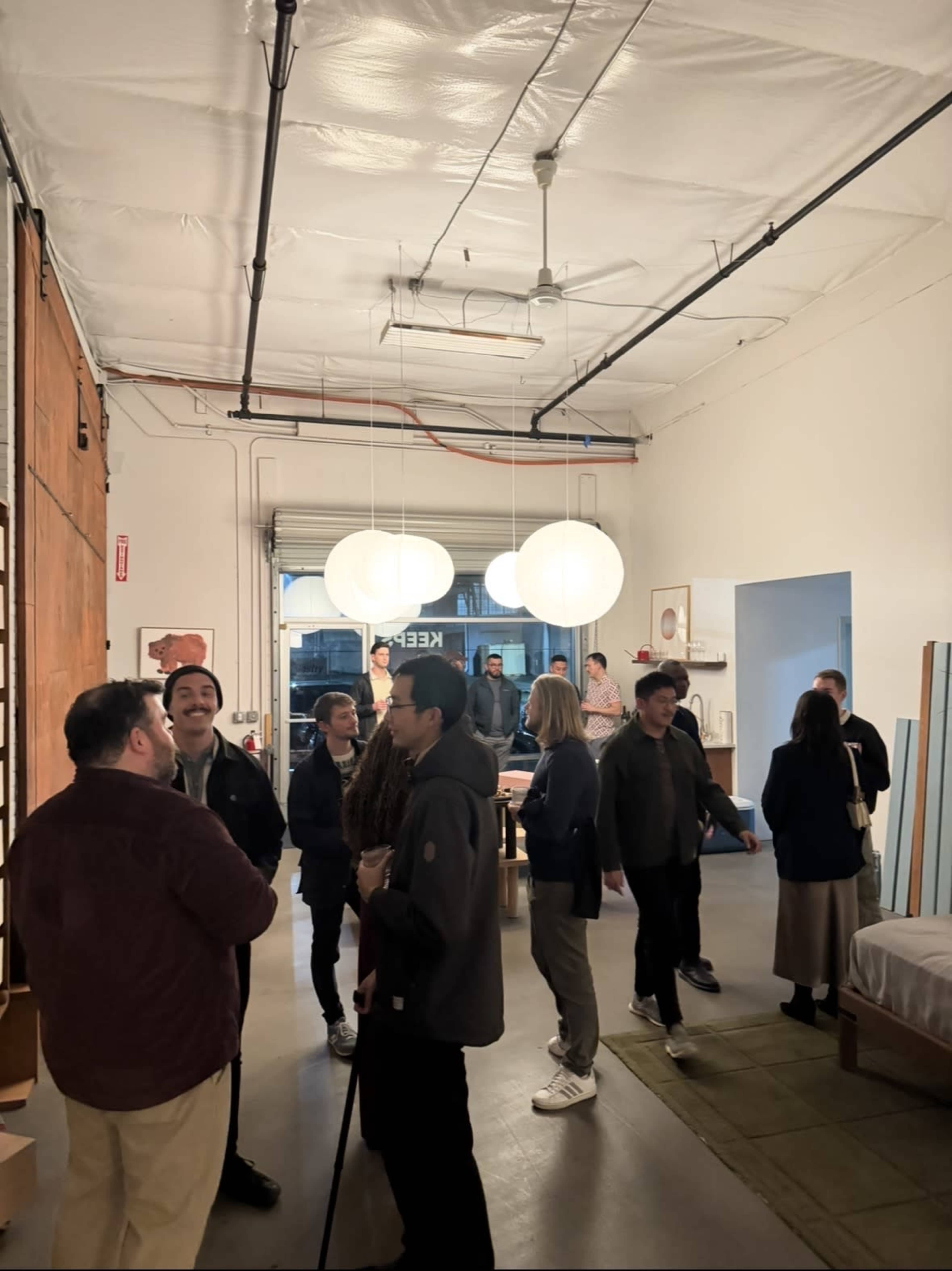 A group of people is gathered in a well-lit, spacious room featuring a mix of furniture items and large pendant lights overhead.