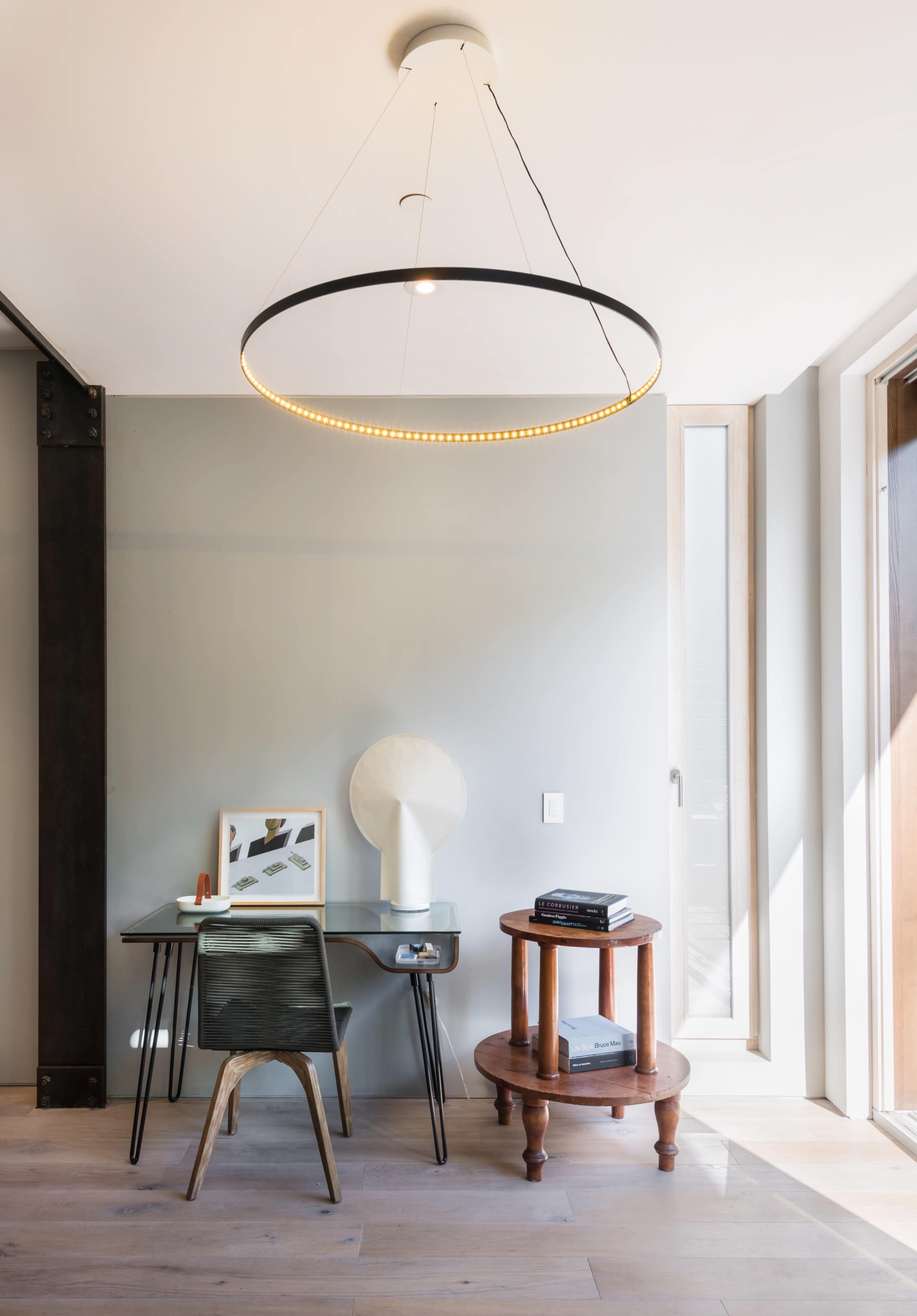A modern workspace features a round pendant light above a desk with a decorative lamp and artwork, alongside a circular wooden side table.
