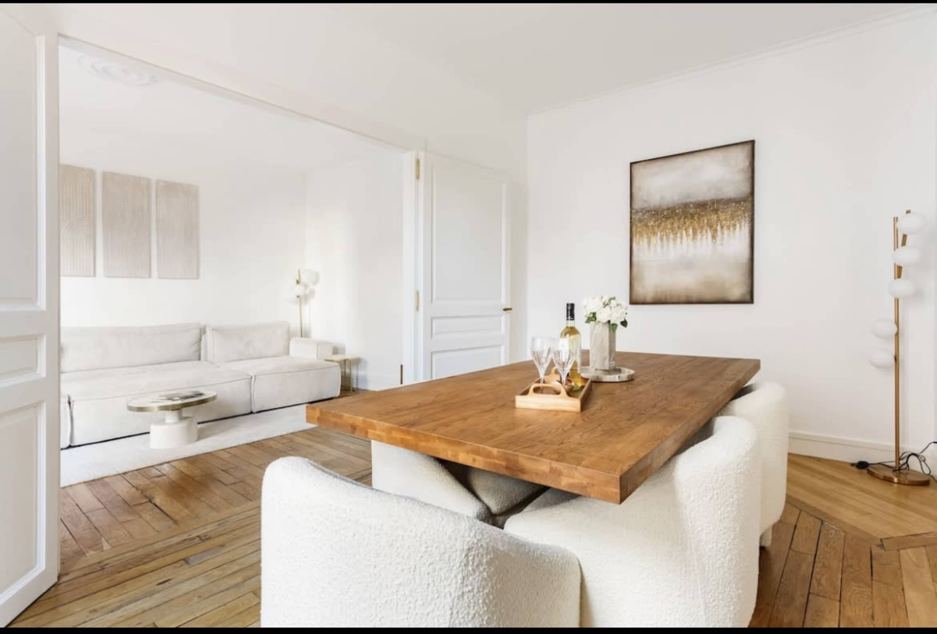 A modern dining area featuring a wooden table surrounded by white, fluffy chairs, with a light-filled living space visible in the background.