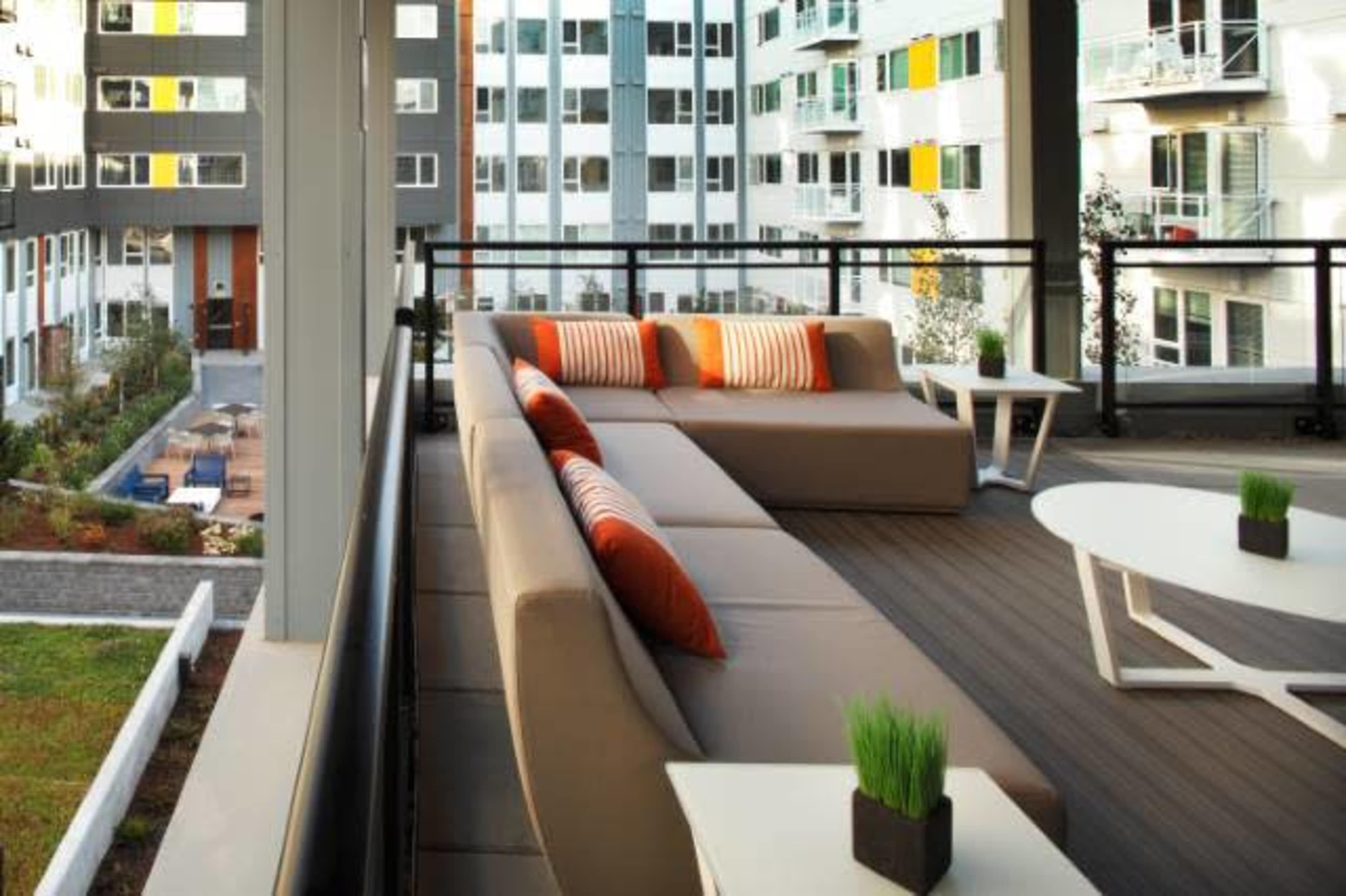 A modern balcony with a large sectional sofa and small tables, overlooking an apartment complex with greenery and a swimming pool below.