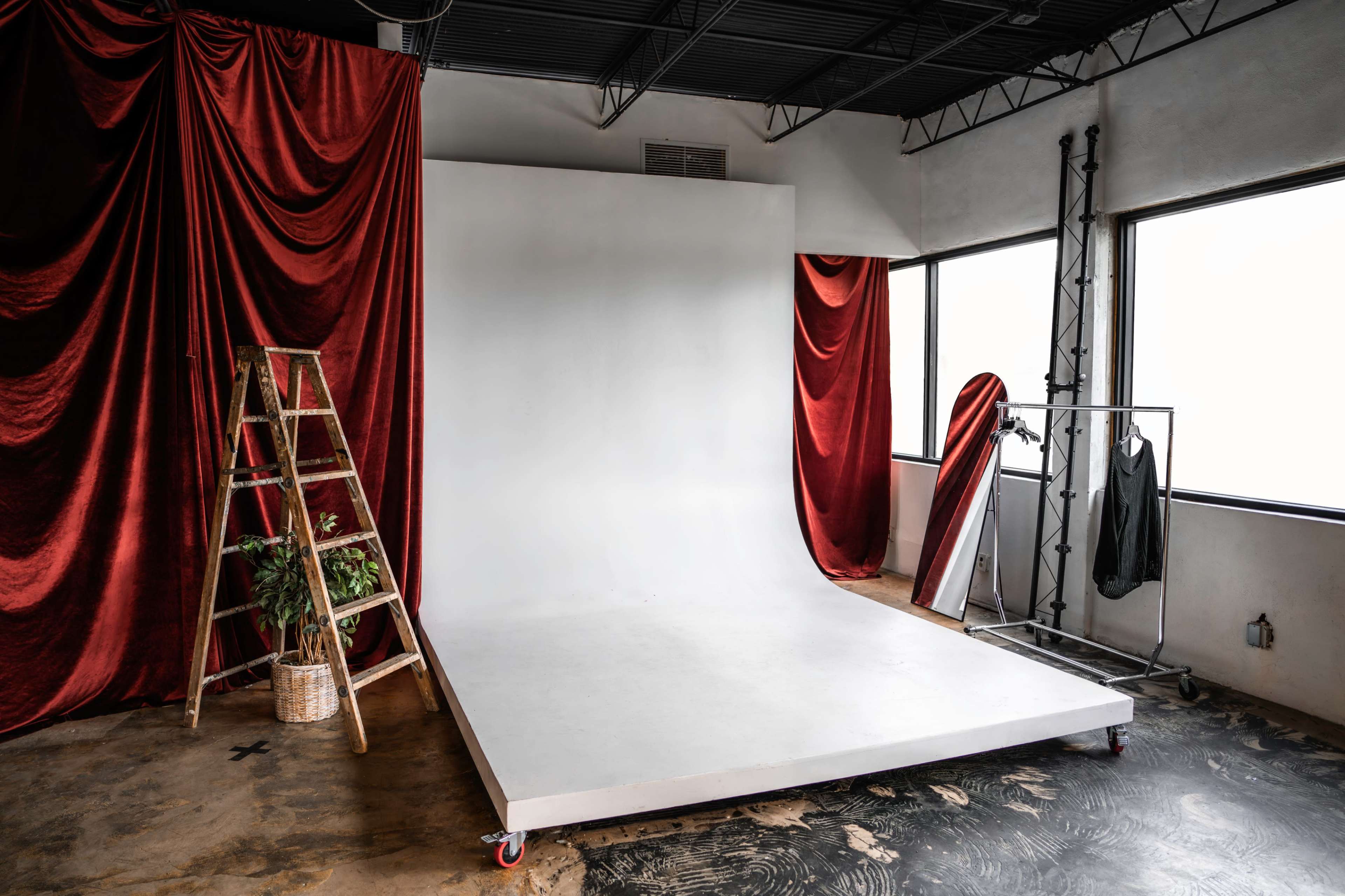 The image shows a photography studio with a white backdrop, a wooden ladder, a clothing rack, and red curtains framing the windows.