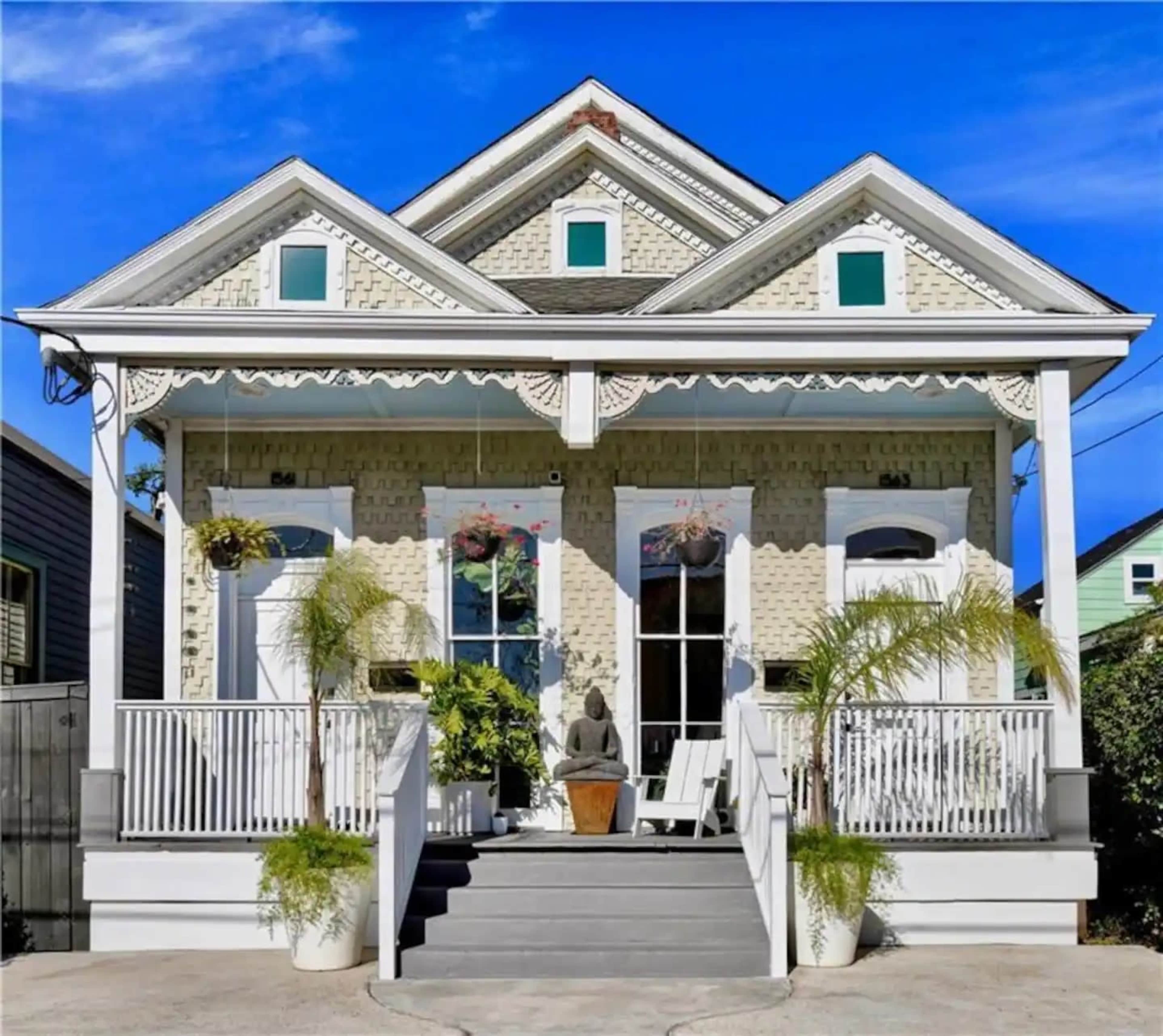 The image shows a symmetrical, two-story house with a light-colored facade, a front porch with decorative railings, and potted plants on either side of the entrance.