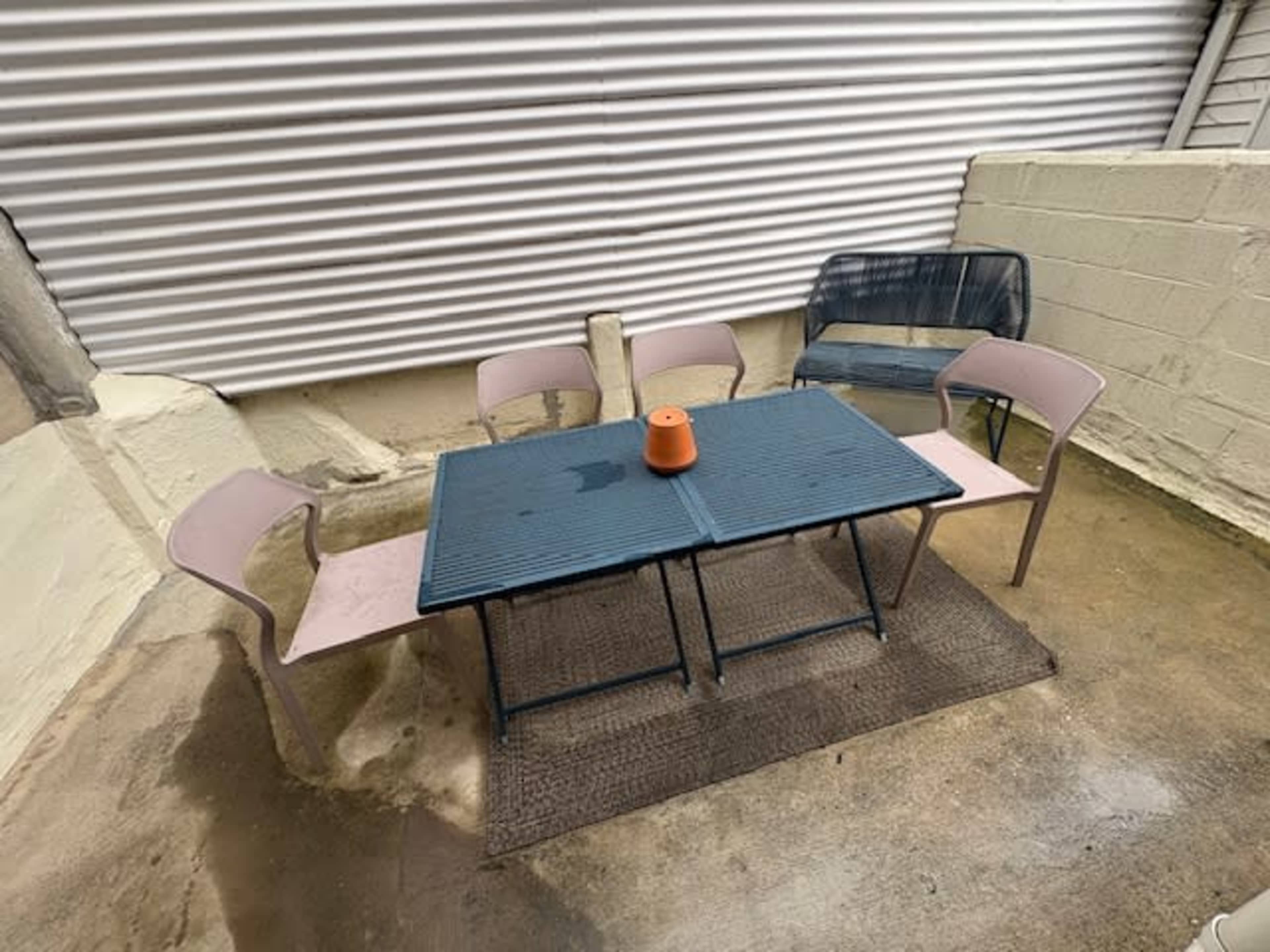 A metal table with a small orange planter is surrounded by four pink chairs on a concrete patio.
