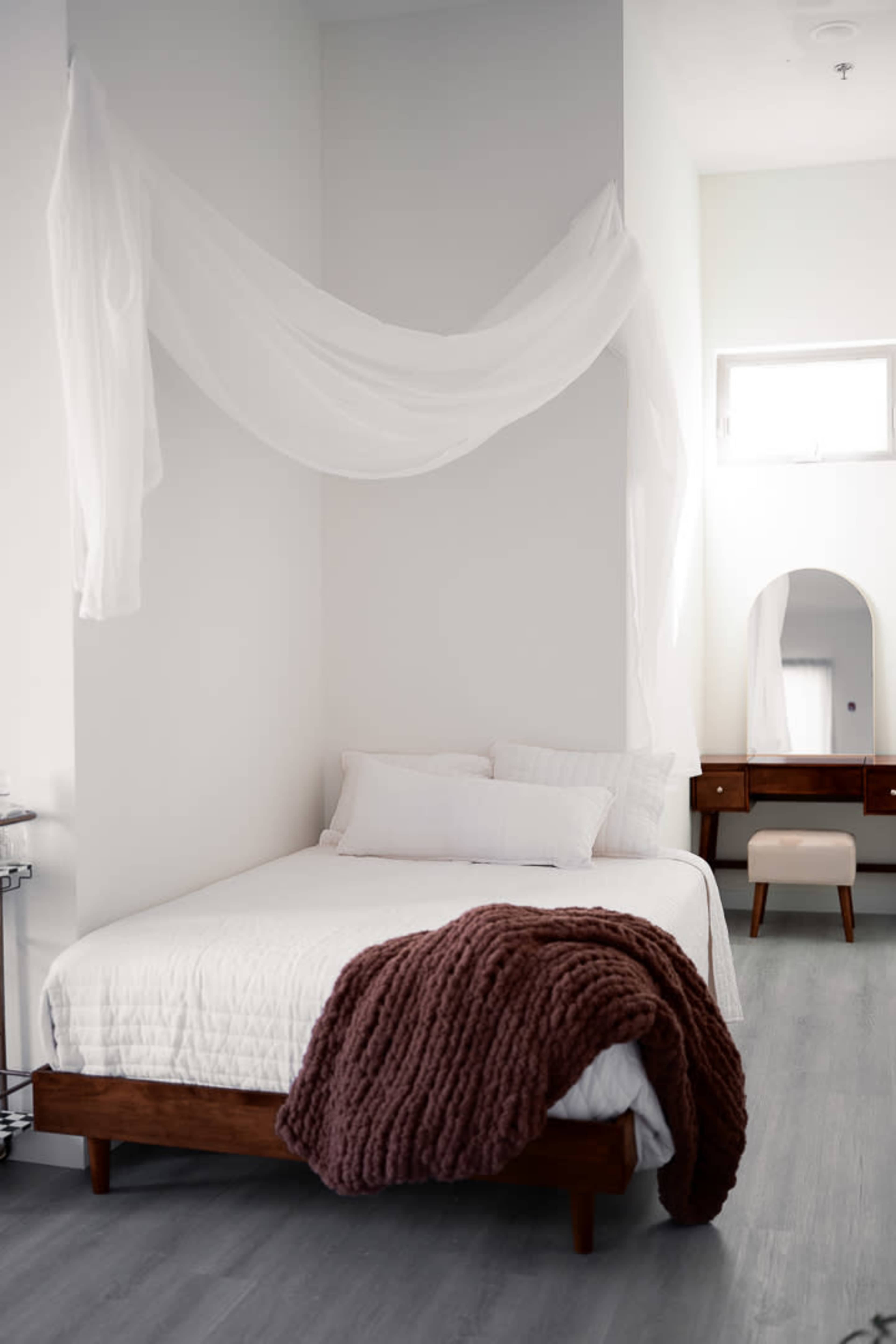The image shows a minimalist bedroom featuring a wooden bed with a white quilt, a brown throw blanket, and sheer white fabric draping above, alongside a wooden vanity with a mirror.