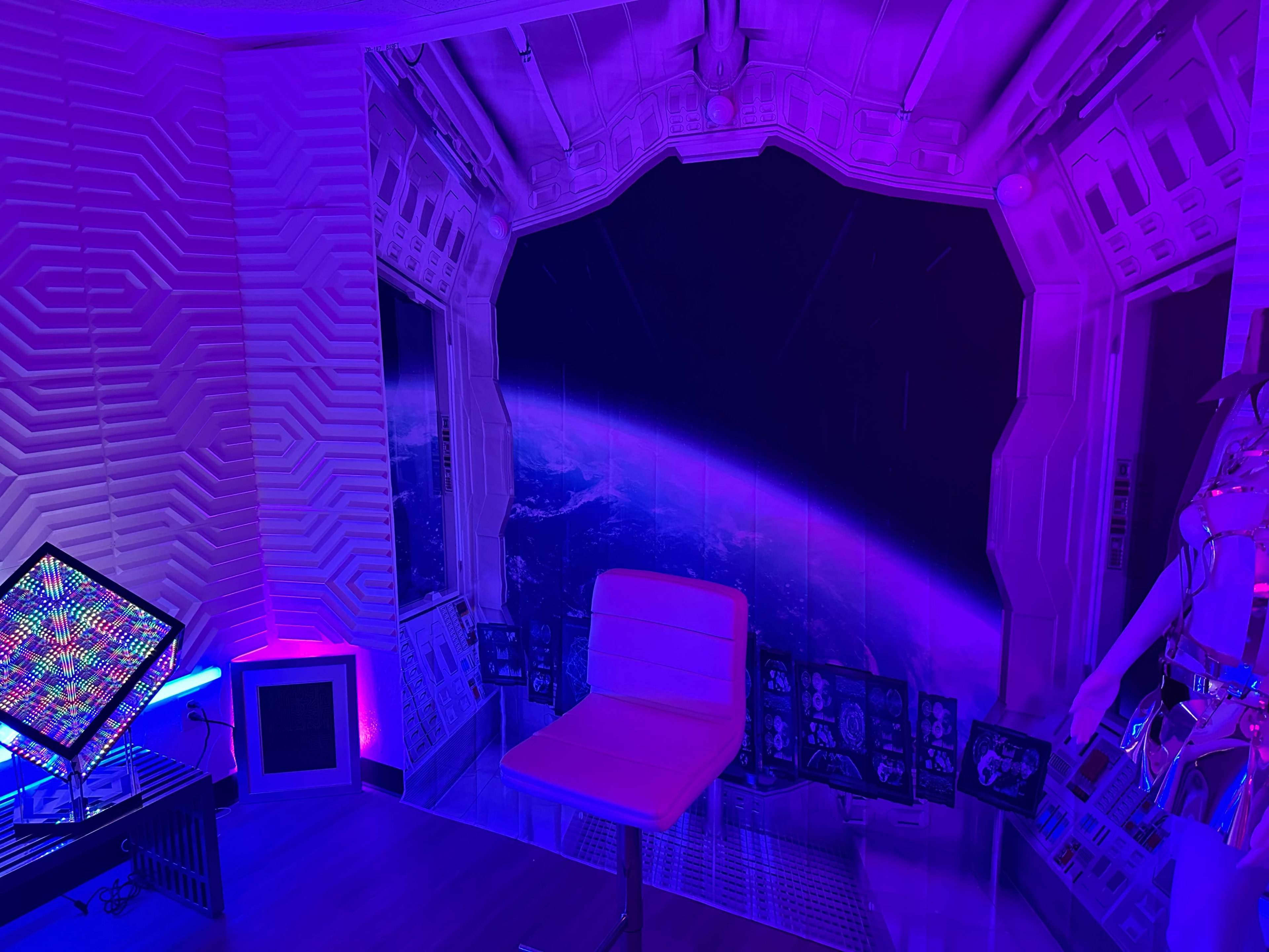 A room with a space-themed wall mural, illuminated by purple lighting, featuring a chair and a colorful light display.