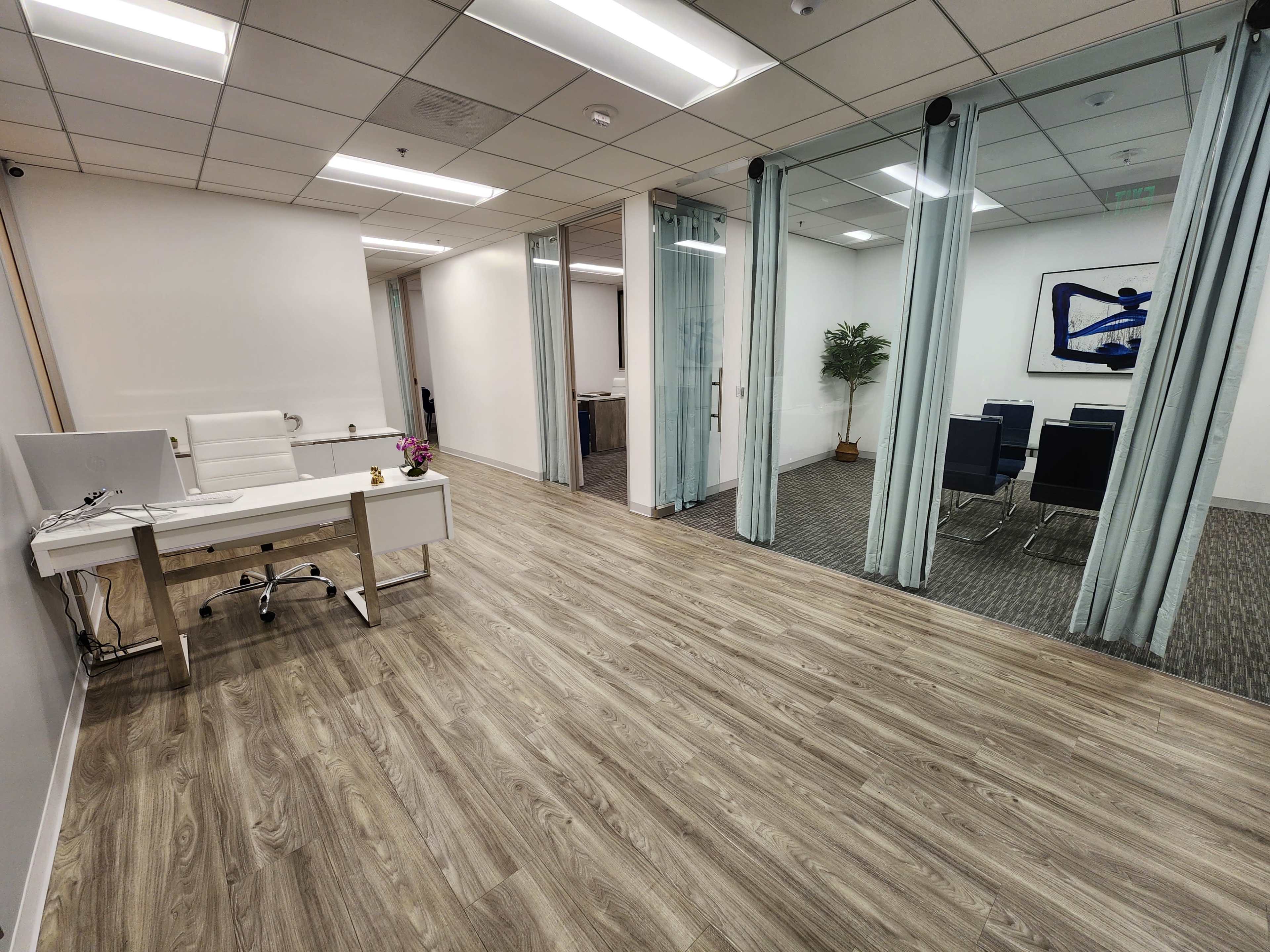 The image shows a modern office space featuring a desk with a computer, a chair, and a meeting area separated by glass partitions.