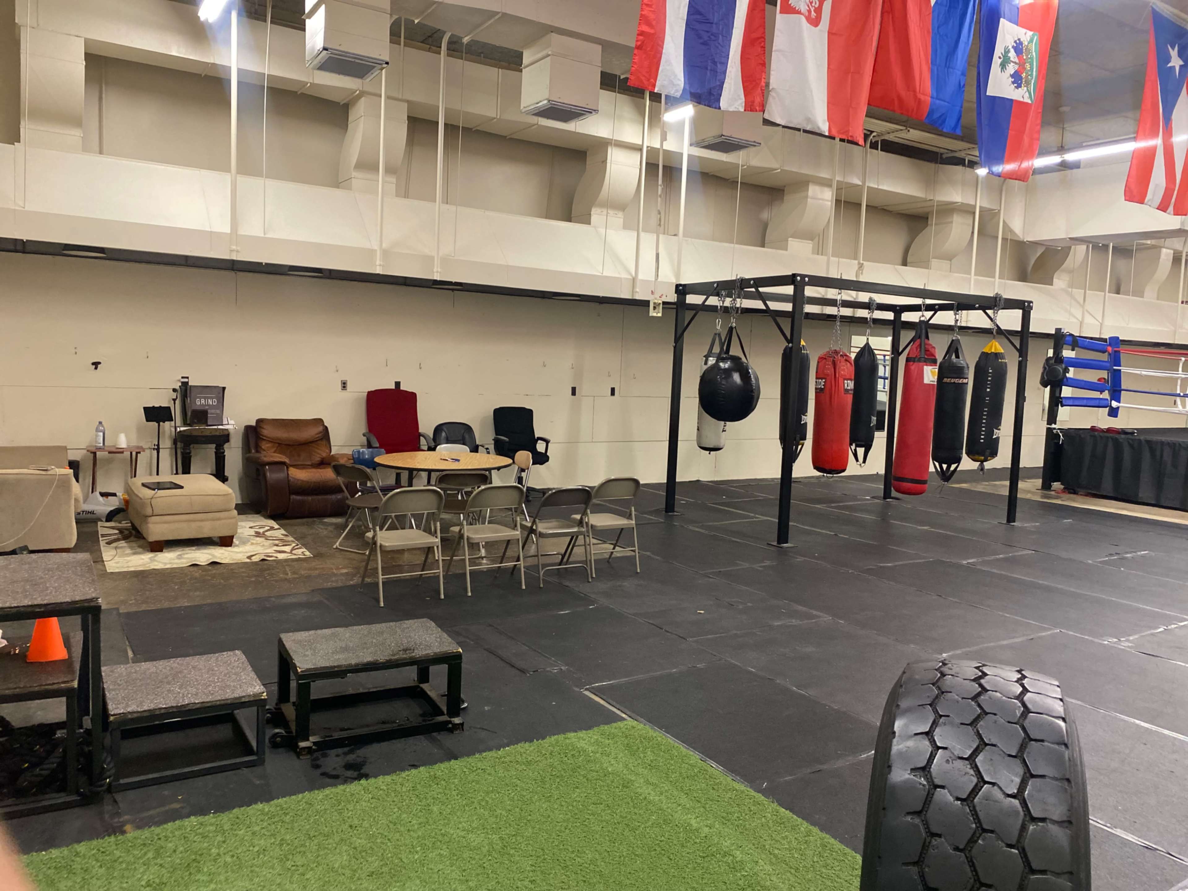 The image shows an indoor training facility with various exercise equipment, including punching bags and a boxing ring, along with a seating area and flags hanging from the ceiling.