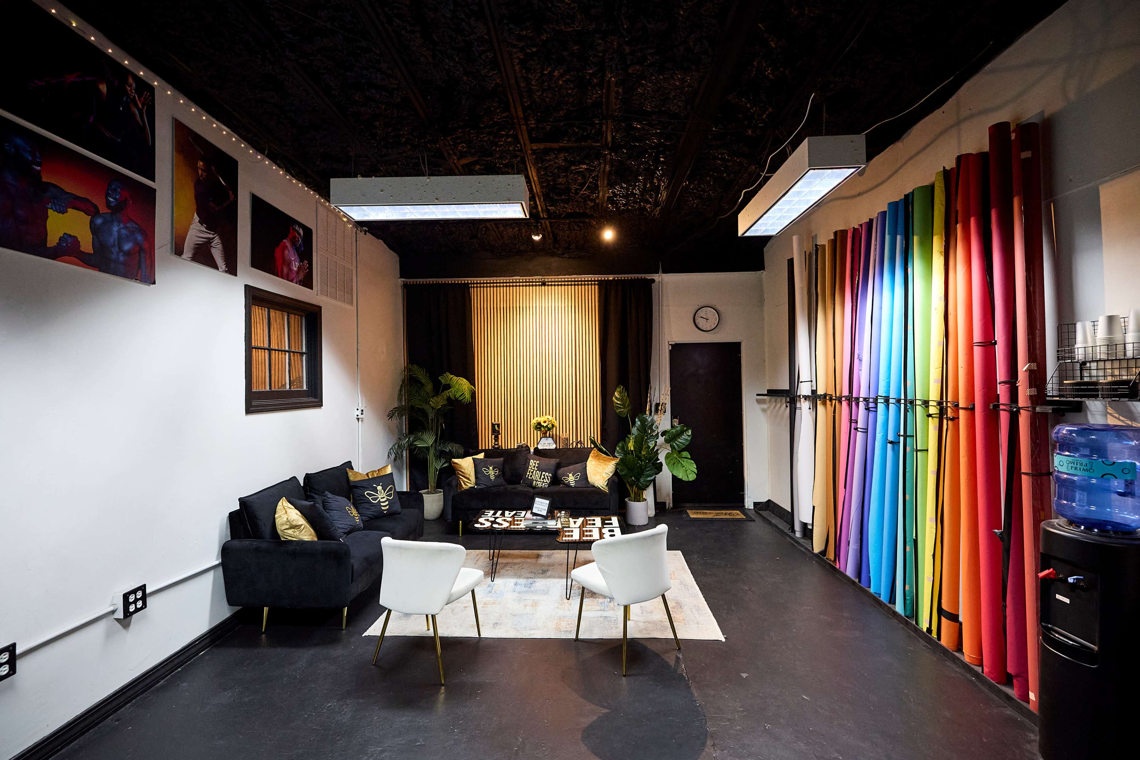 The image shows a spacious studio room featuring a black couch with decorative pillows, a coffee table, and vibrant colored backdrops hanging on the wall.