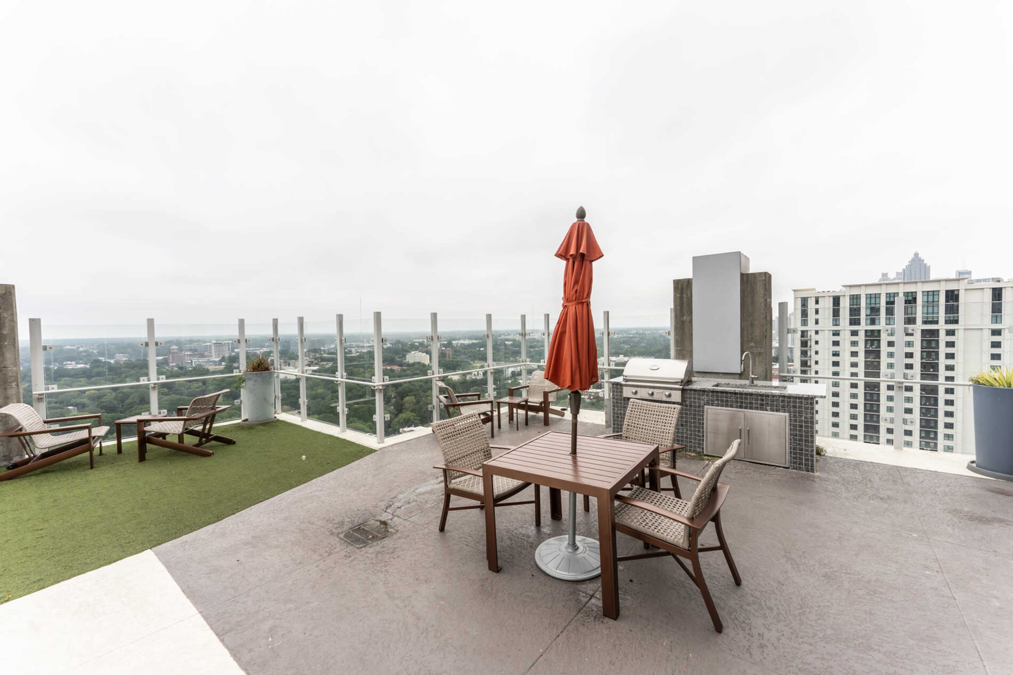 The image shows a rooftop patio area with a dining table, chairs, an orange umbrella, and a grill, surrounded by a railing with a view of a cityscape.