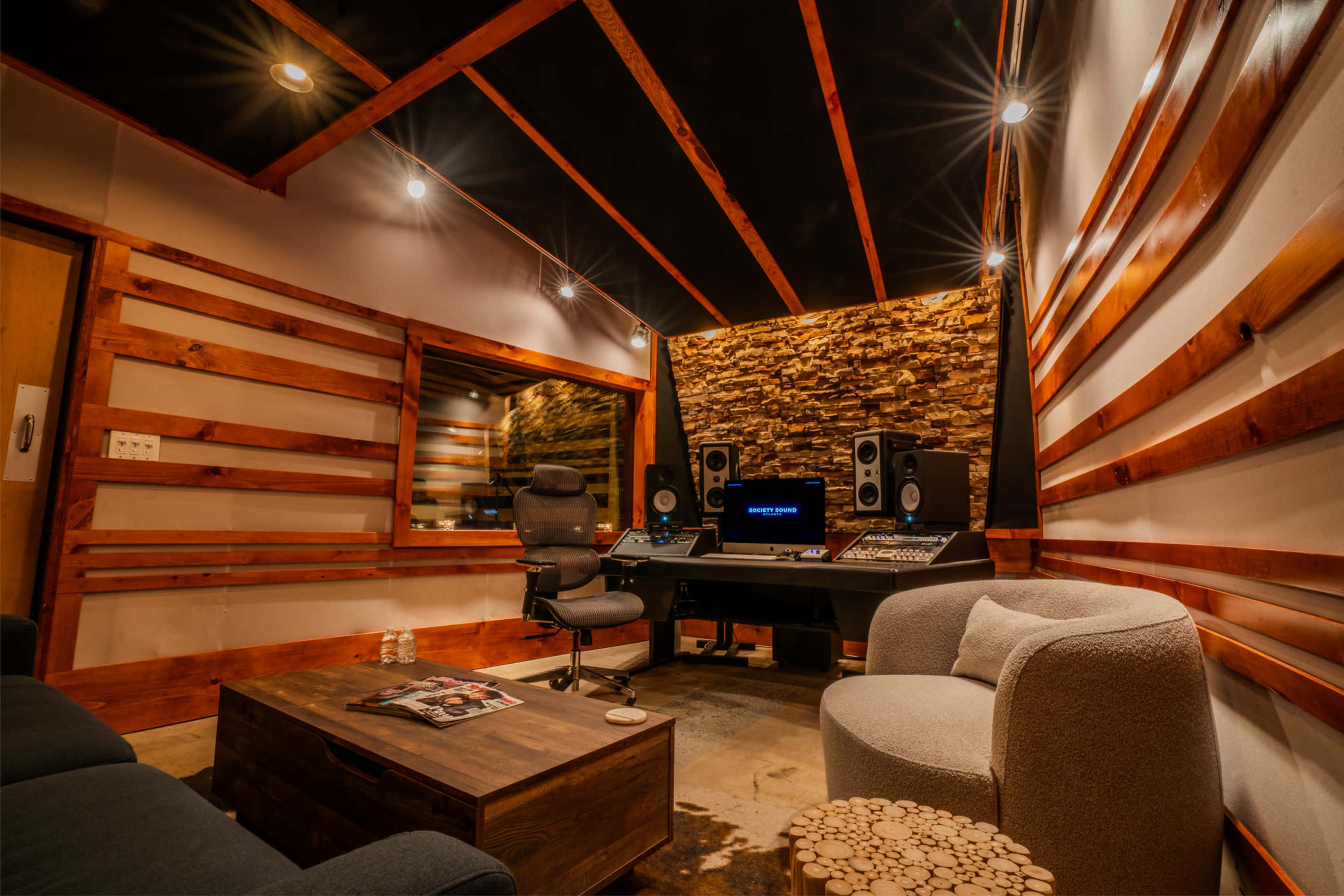 The image shows a modern recording studio featuring a central mixing console, two large speakers, a comfortable chair, and a coffee table surrounded by wooden wall accents.
