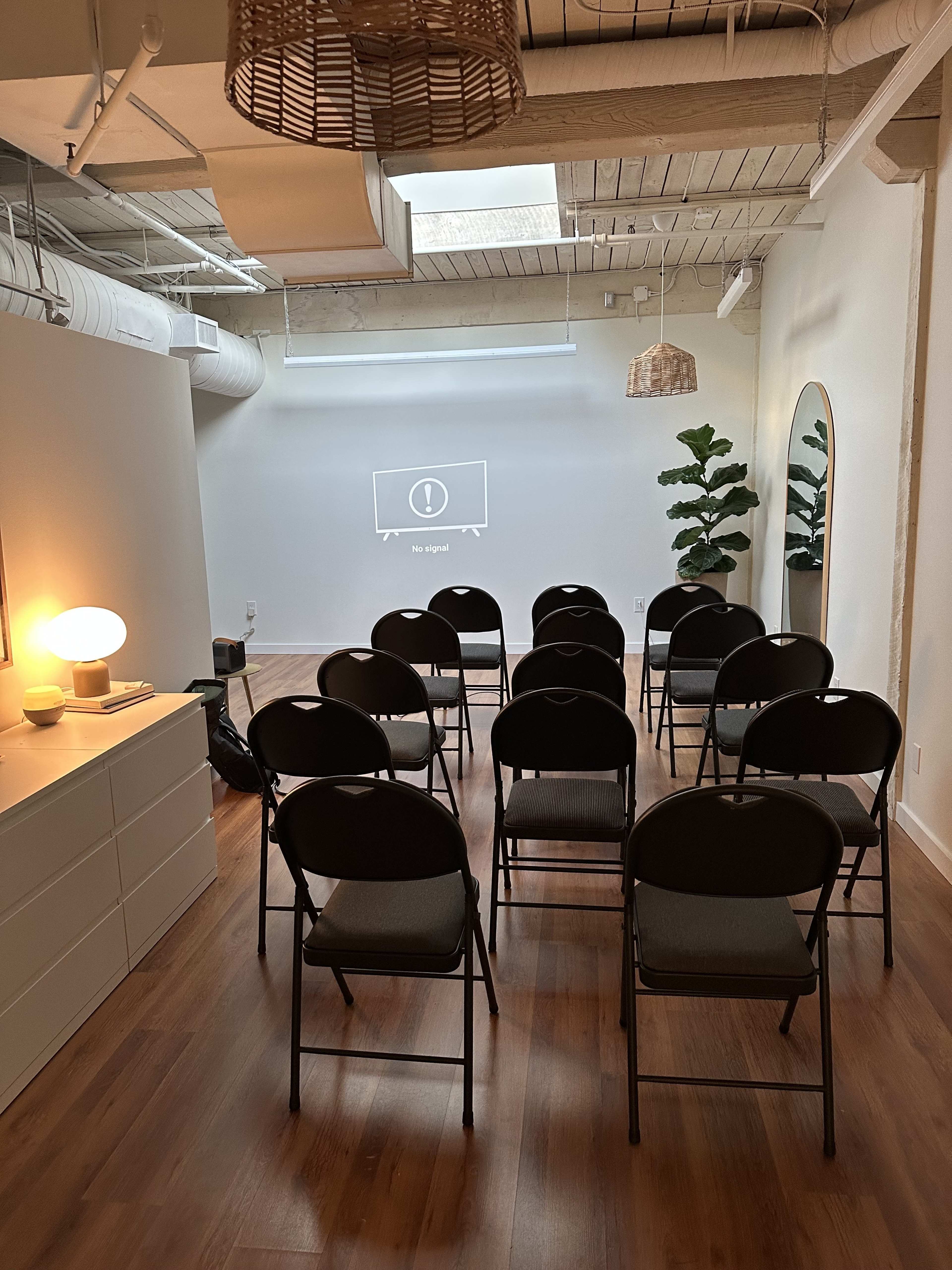A presentation room features rows of black folding chairs facing a blank projector screen, with a modern decor style including potted plants and woven light fixtures.