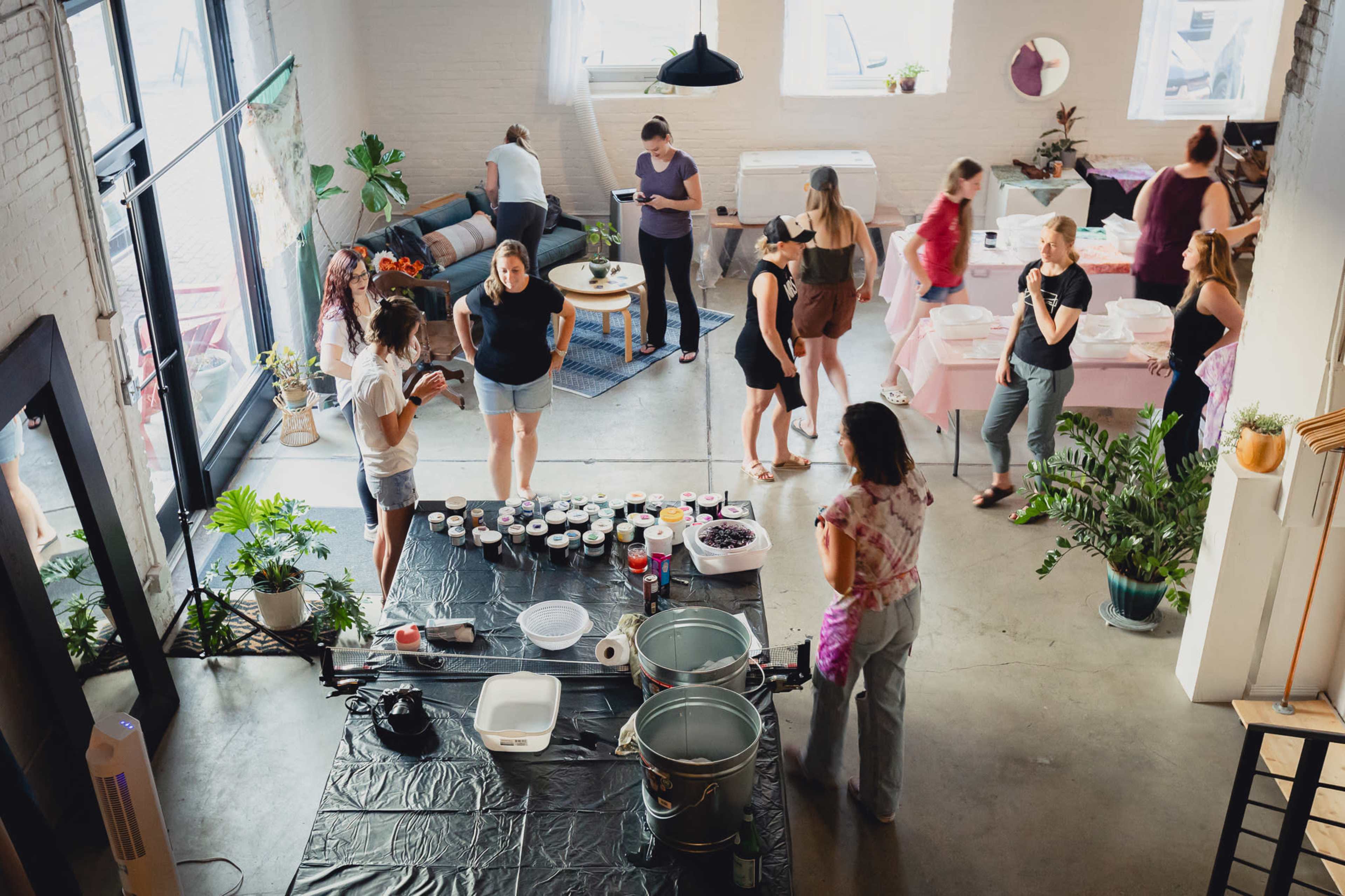 A group of people engages in various activities within a spacious room adorned with plants and tables, while supplies for a craft project are organized on a black table.