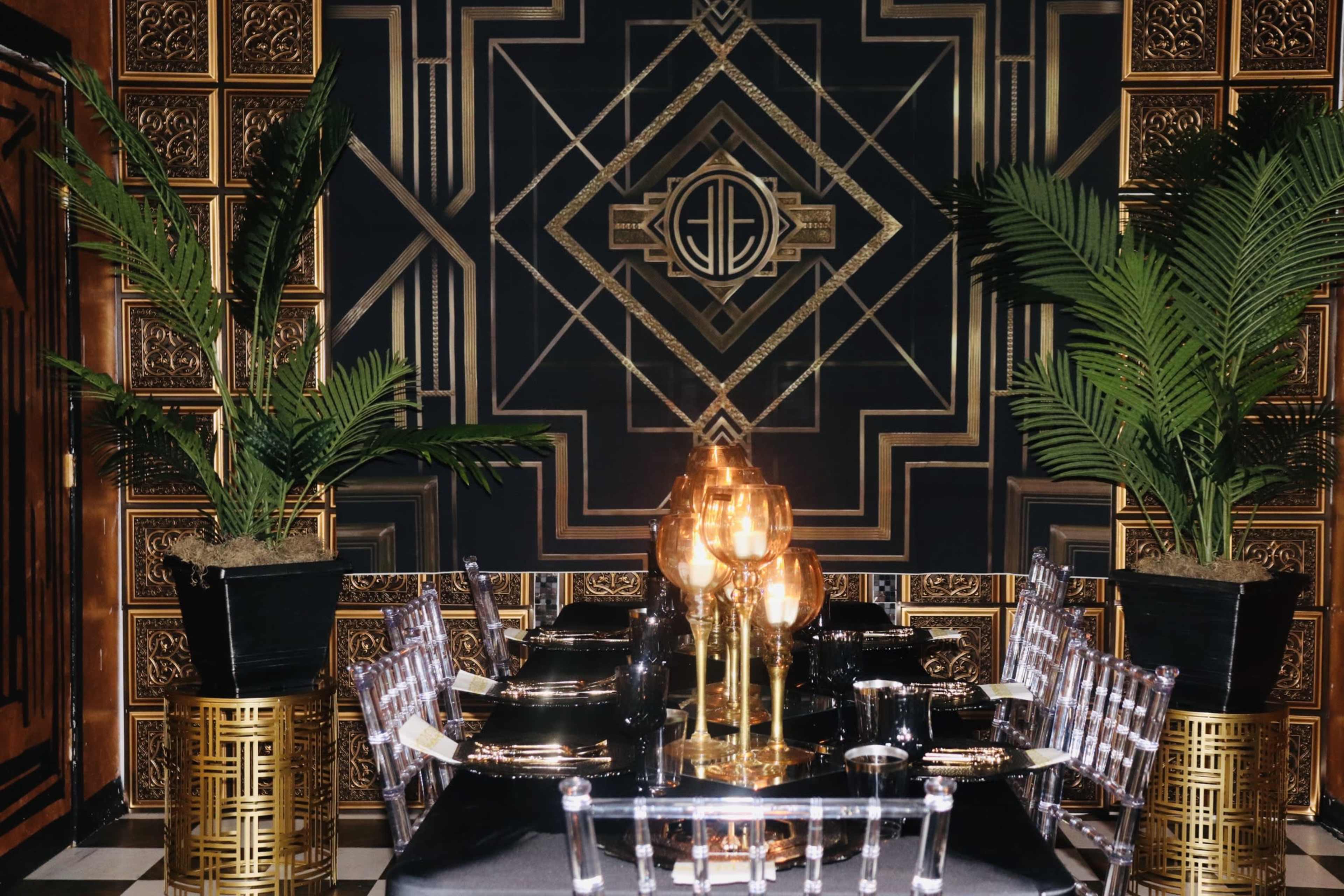 An elegantly designed dining room featuring a black and gold geometric wall pattern, a glass dining table, and decorative plants in planters.