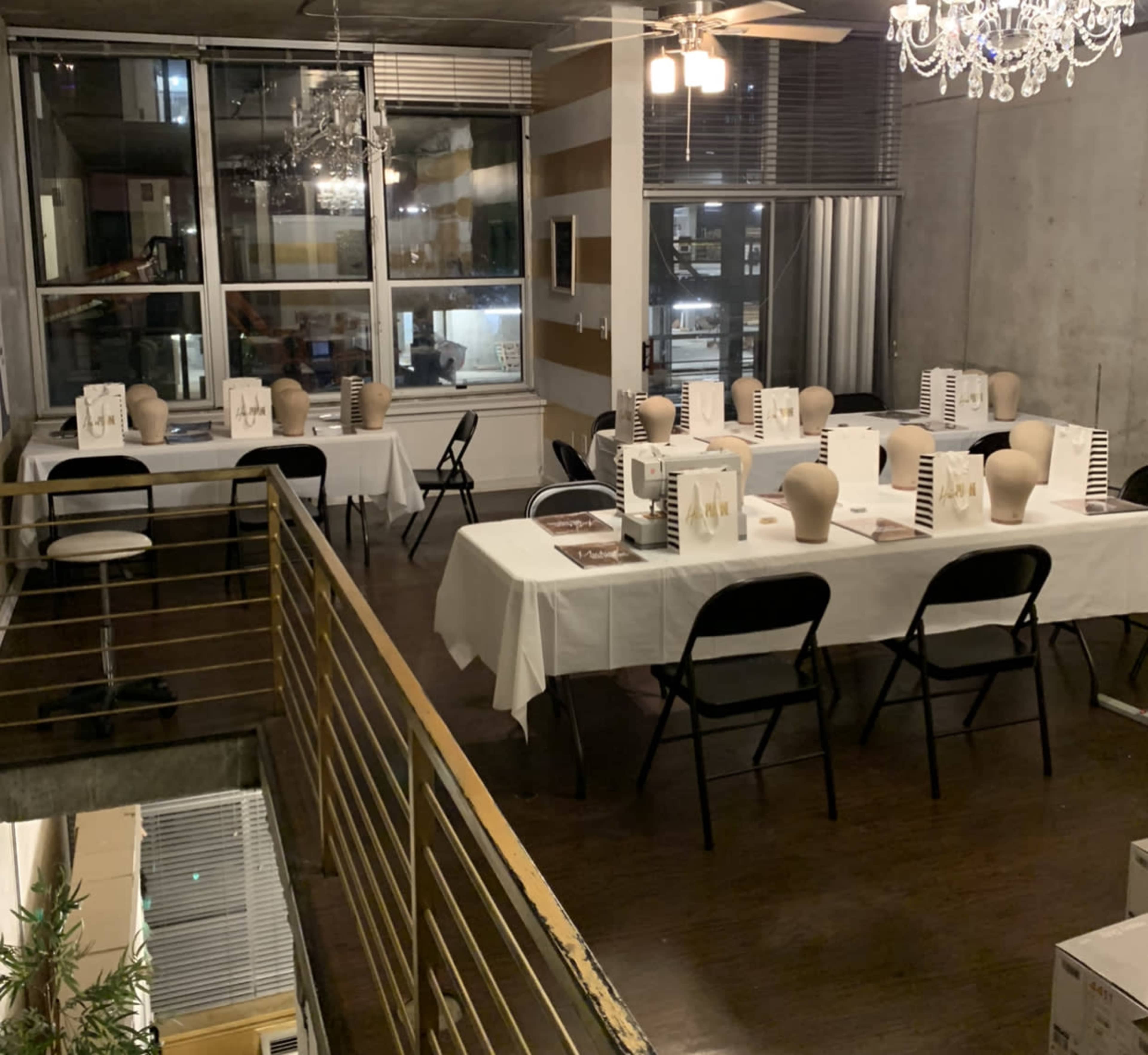 A spacious room with several tables arranged in a U-shape, each covered with white tablecloths and topped with head mannequins, and large windows letting in light.