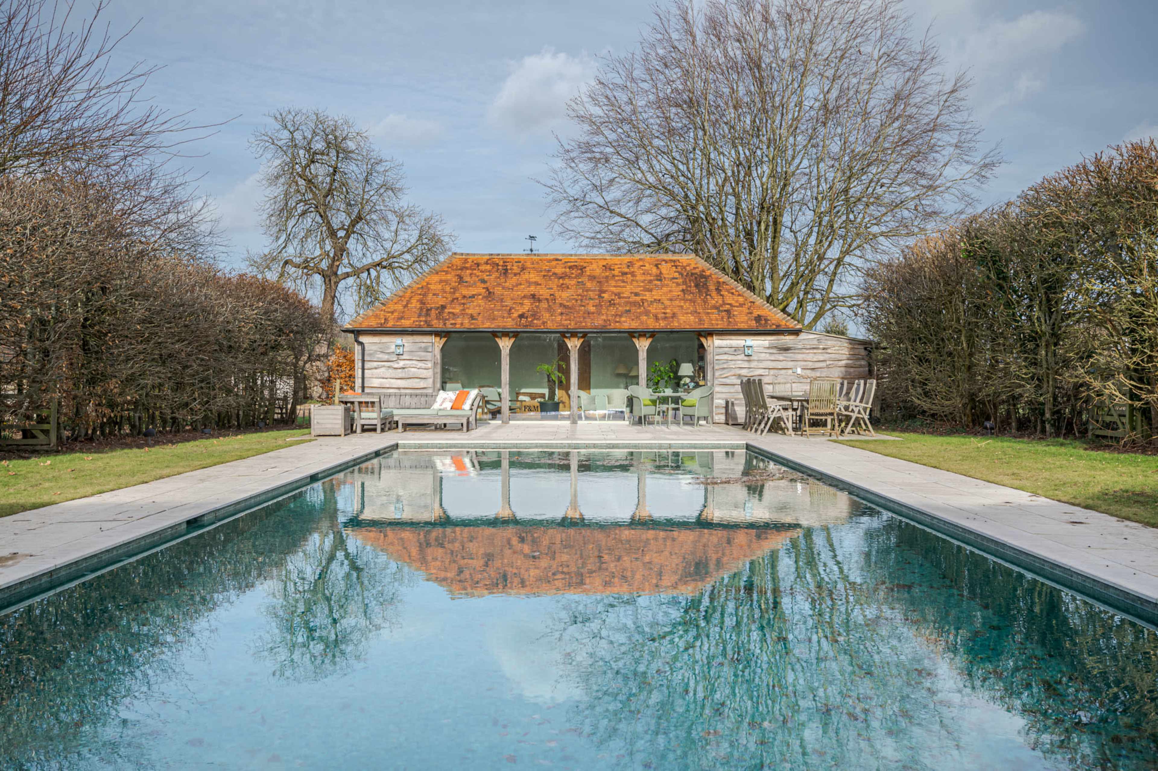 A tranquil outdoor scene features a rectangular swimming pool reflecting a wooden cottage with a rust-colored roof, framed by trees and neatly trimmed hedges.