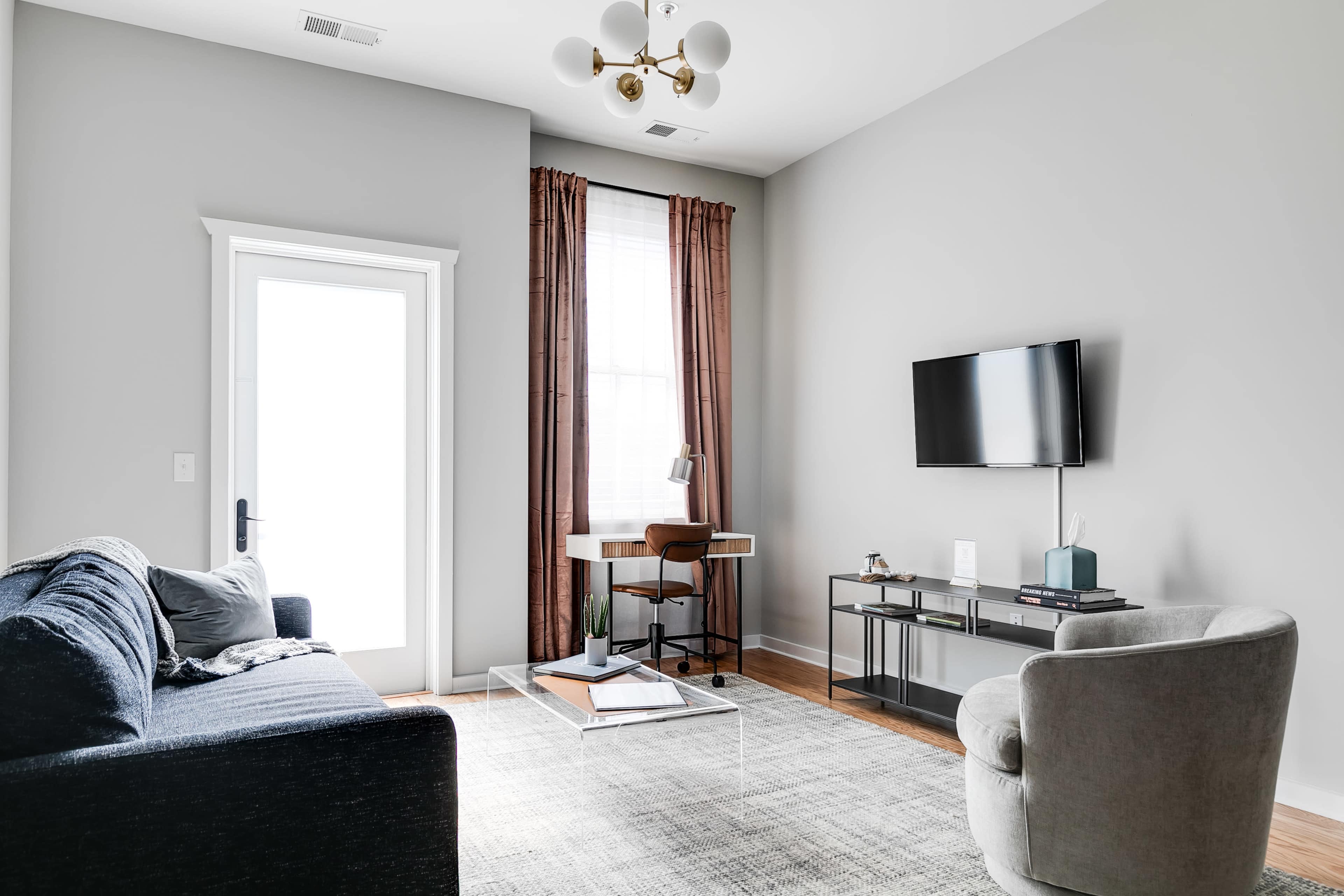 A modern living room with a blue sofa, a round chair, a glass coffee table, a desk with a chair, and a wall-mounted TV.