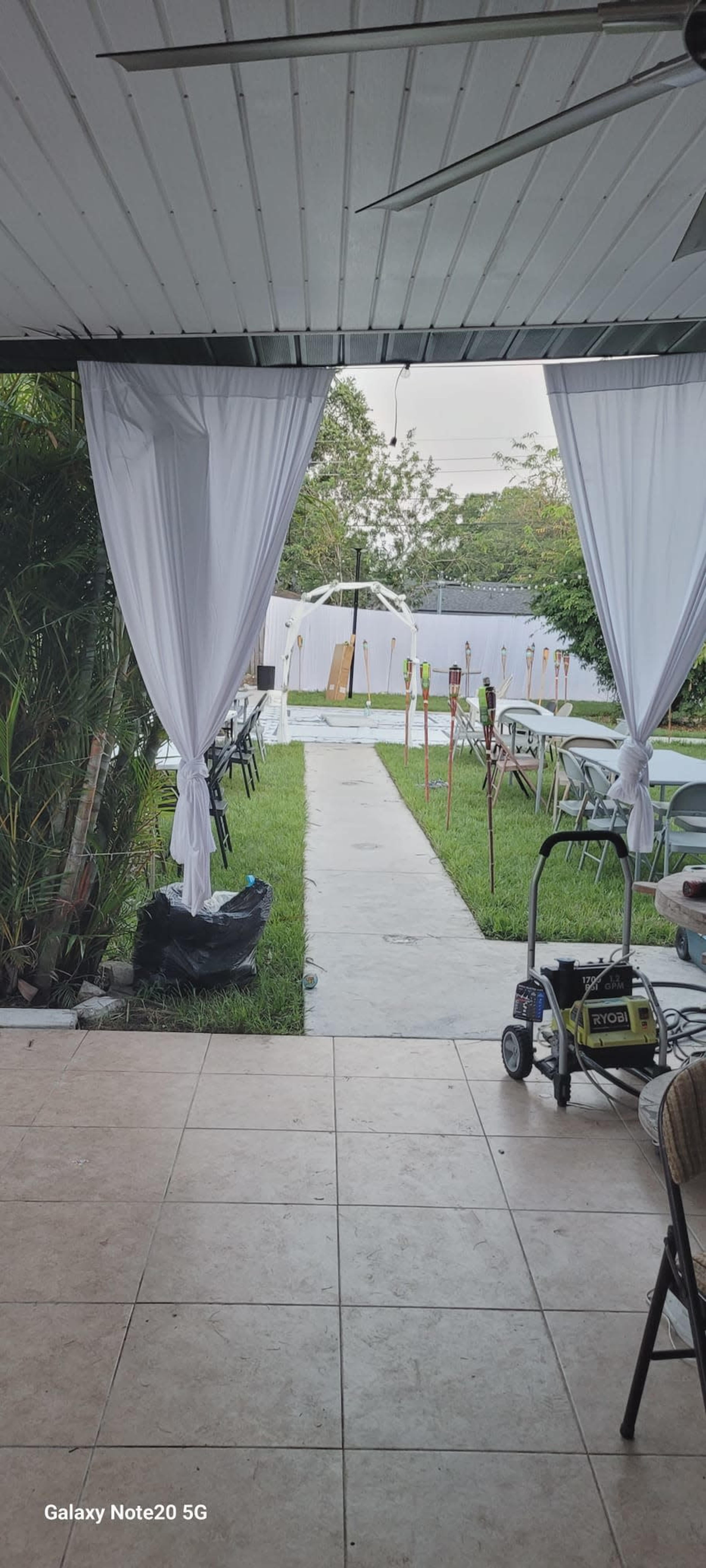 The image shows an outdoor space with a pathway lined by grass and tables set up for an event, viewed from a covered area with draped curtains.