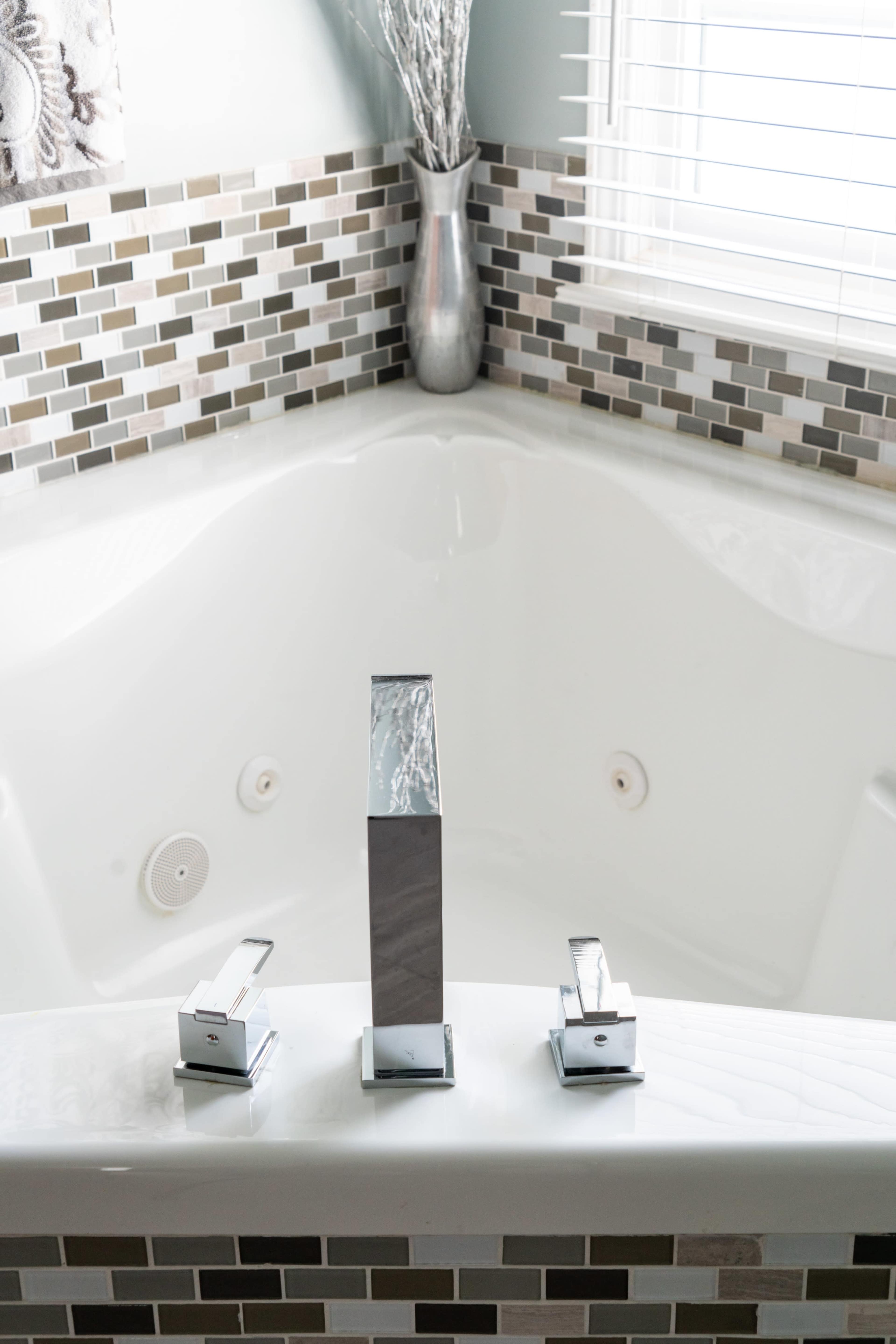 The image shows a modern bathtub with three chrome faucets and a mosaic tile backsplash.