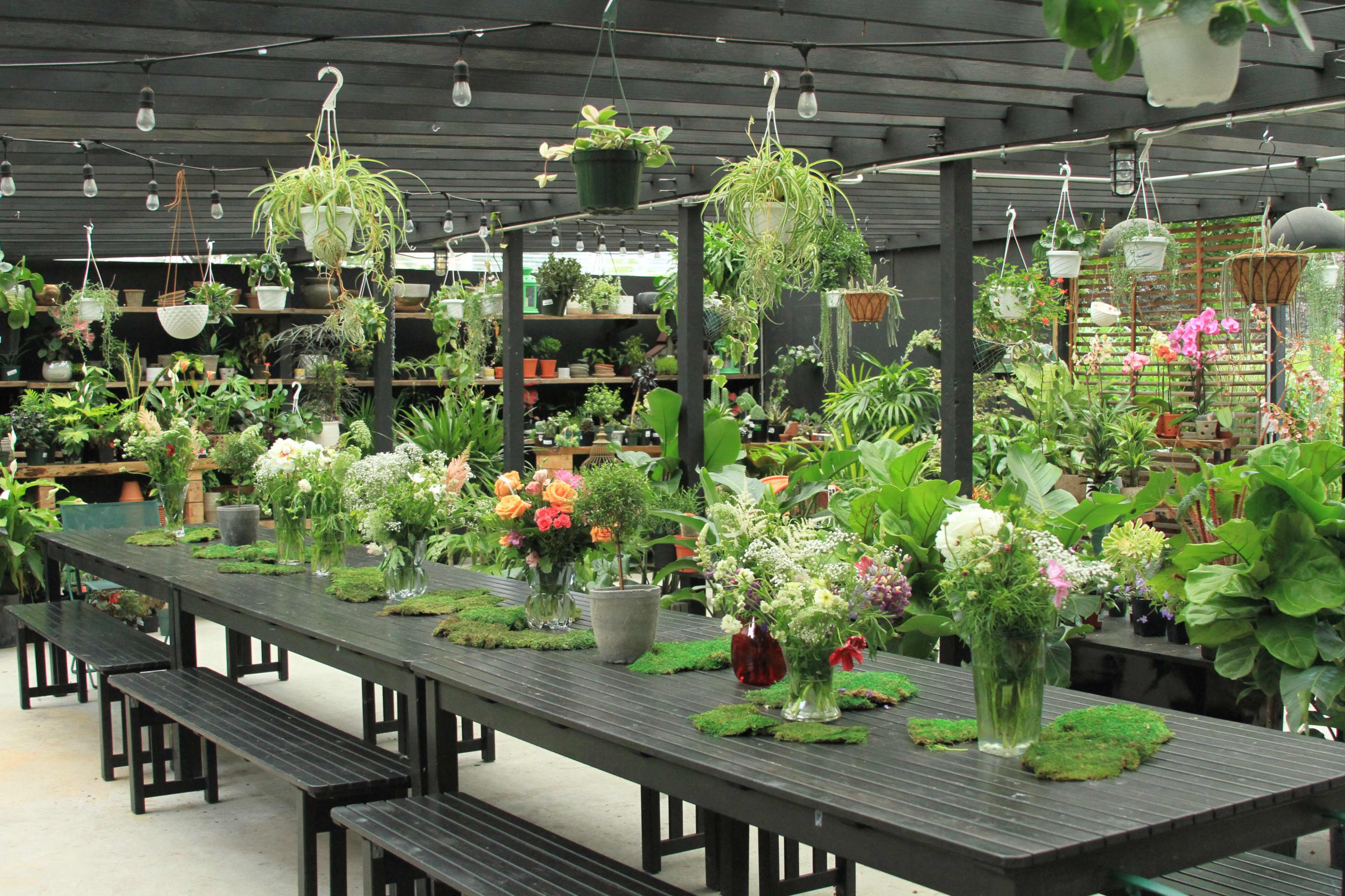 The image shows a greenhouse filled with various plants and flowers, featuring a long black table adorned with vases of fresh flowers and greenery.