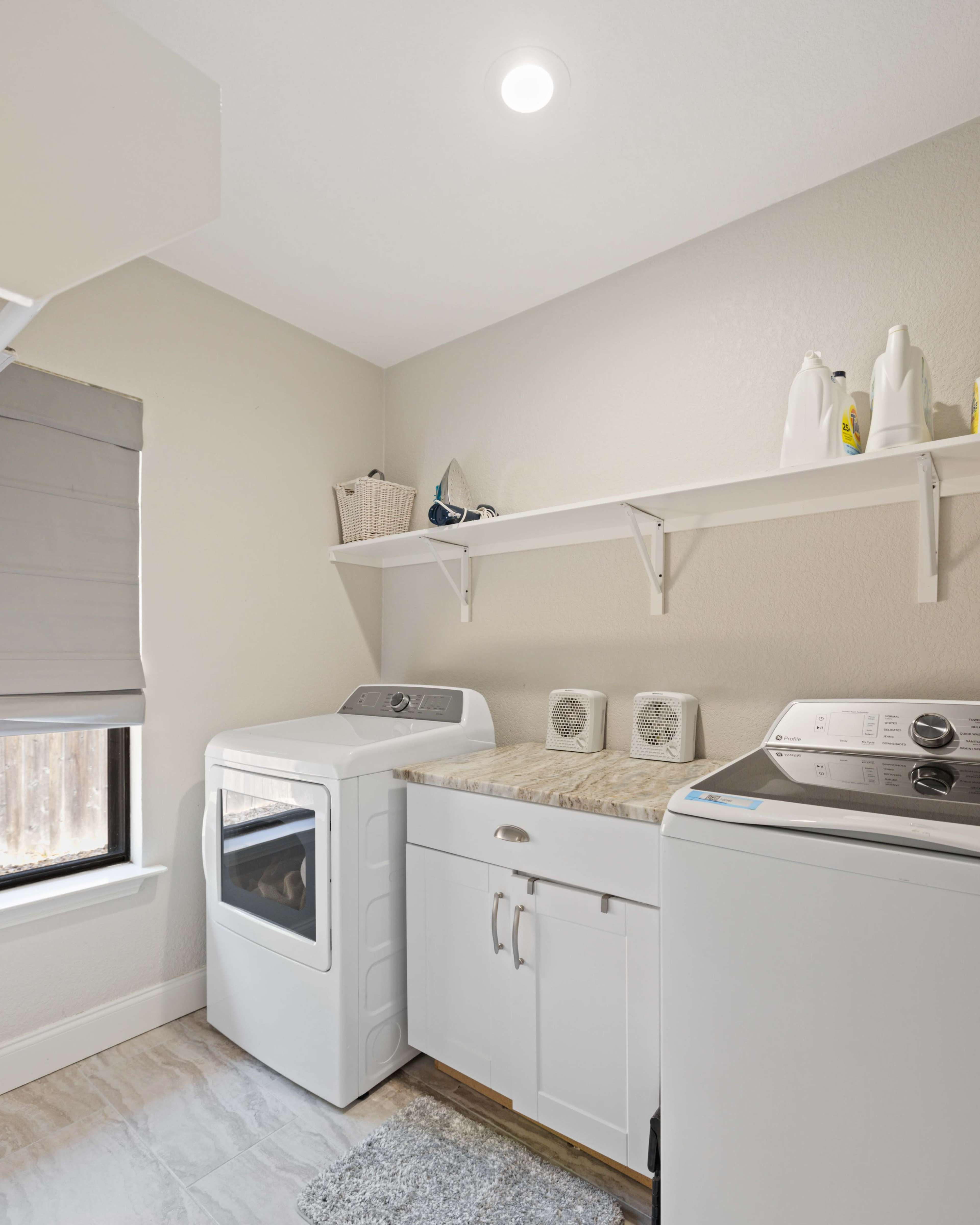 A laundry room features a stacked washer and dryer beside a countertop with shelves above for storage.