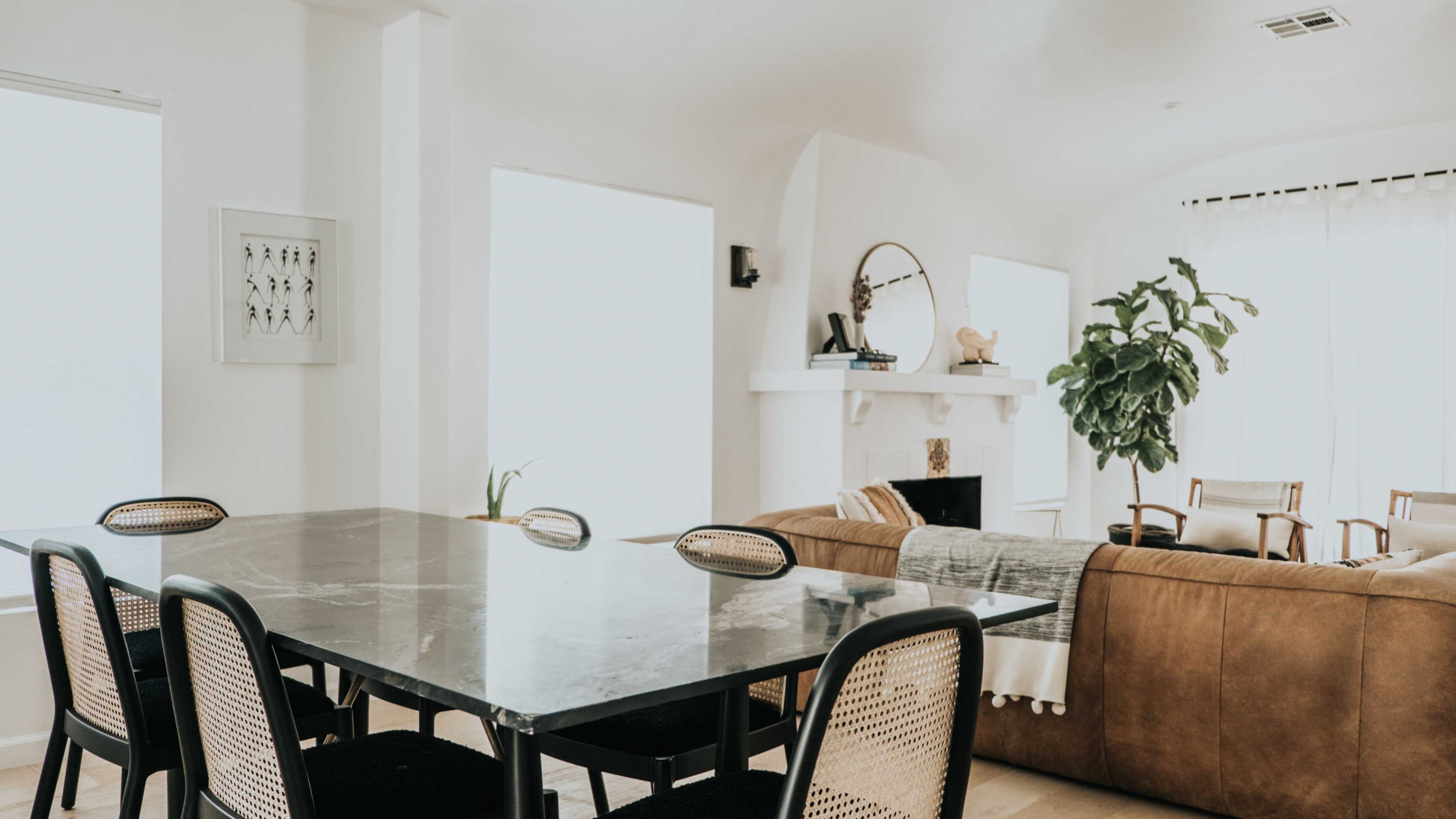 The image shows a modern living space featuring a dining table with black chairs and a brown sofa, along with large windows and potted plants.