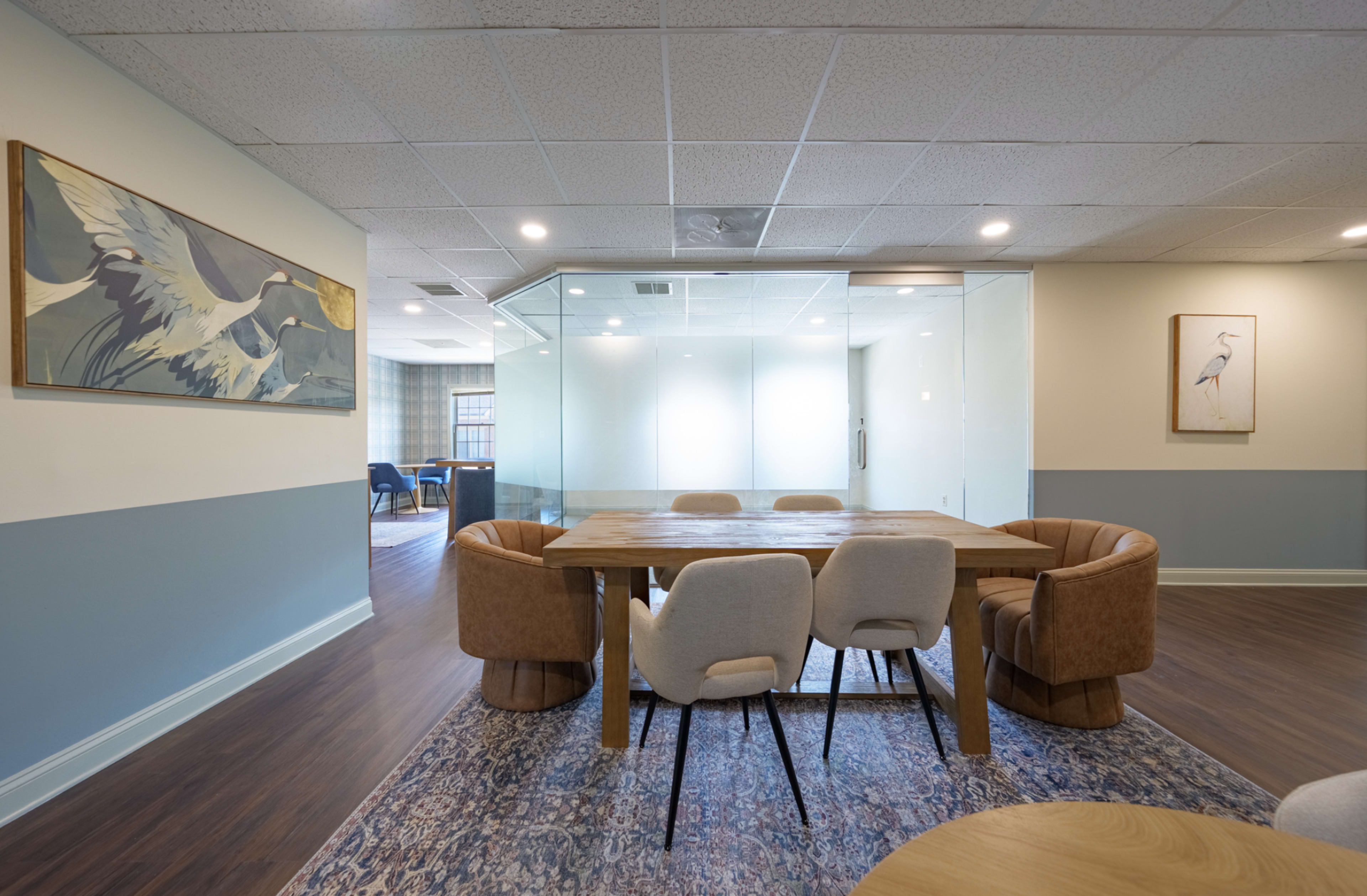 The image shows a modern office space with a wooden table surrounded by upholstered chairs, a glass conference room enclosed in a partition, and artwork on the walls.