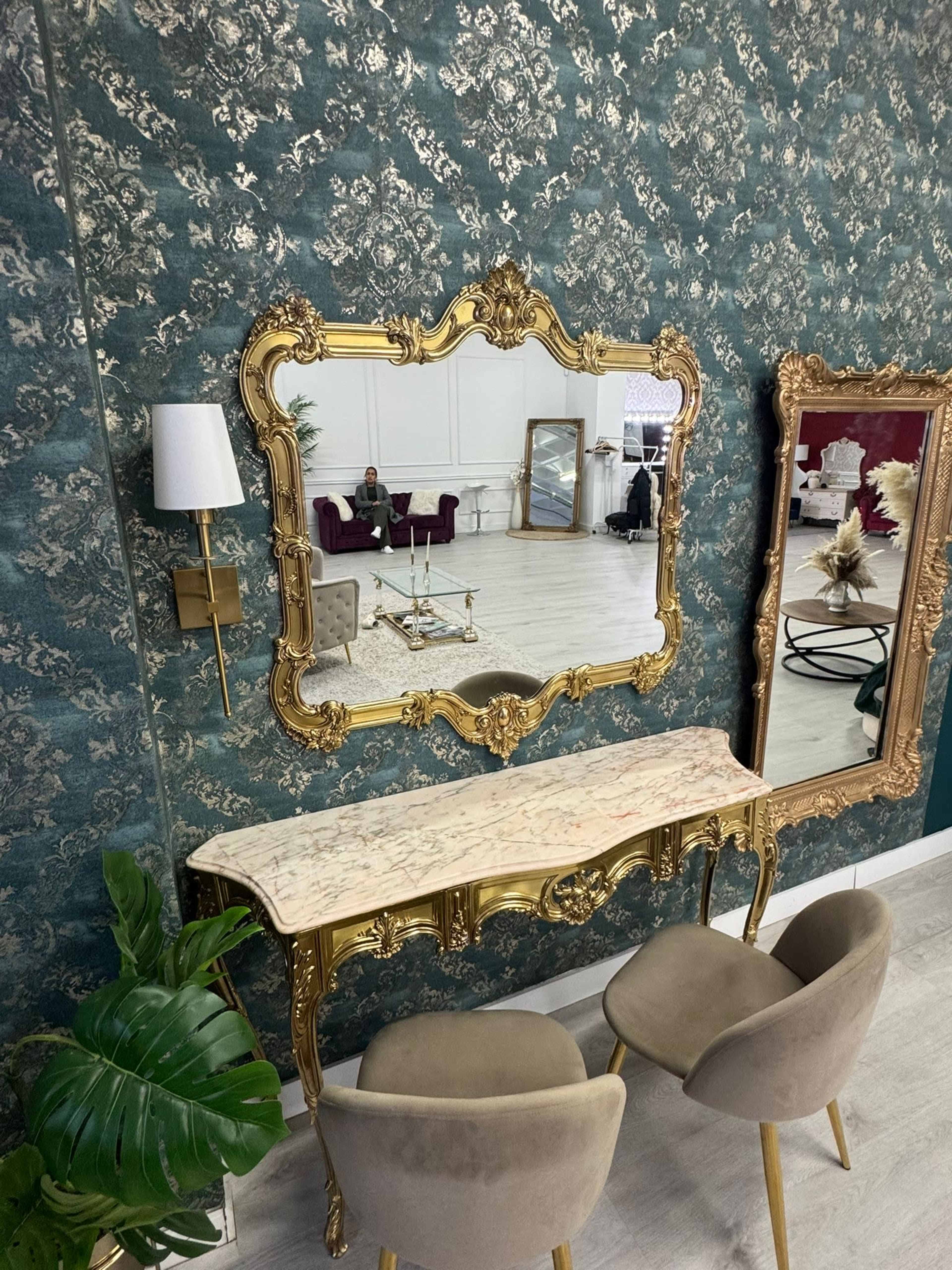 The image features an ornate gold-framed mirror above a marble-topped console table, with two upholstered chairs and a decorative wall exhibiting a patterned design.