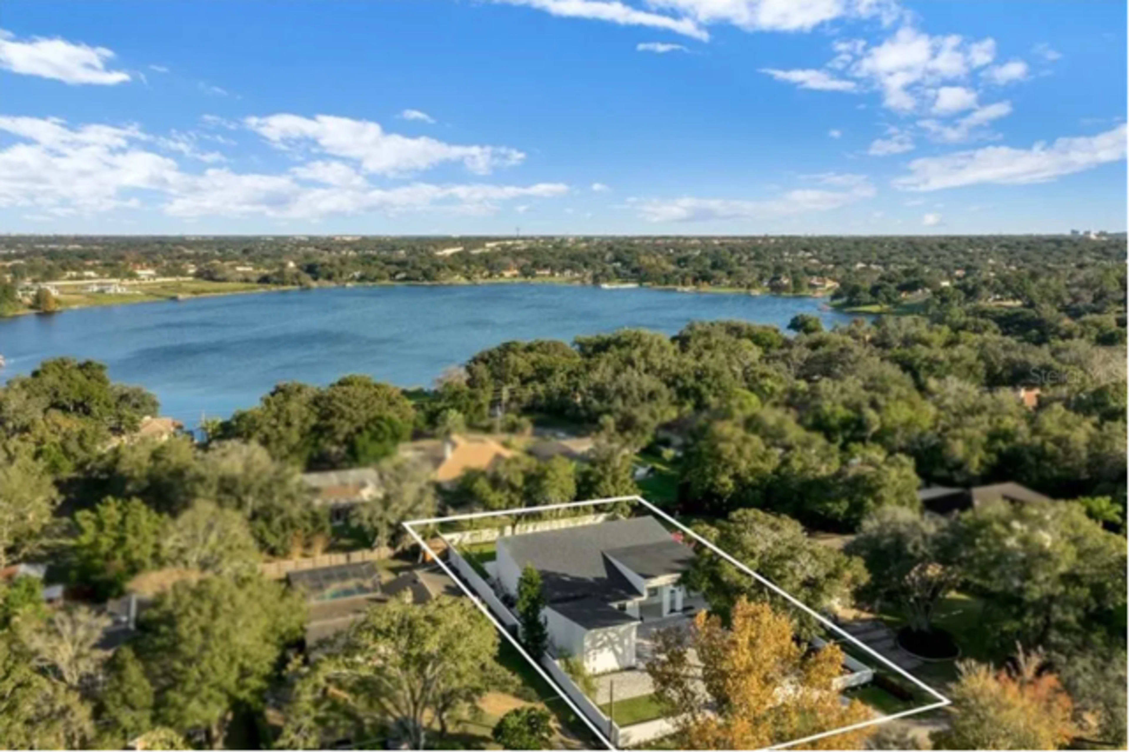 The image shows an aerial view of a house surrounded by trees, situated near a lake with a clear blue sky above.