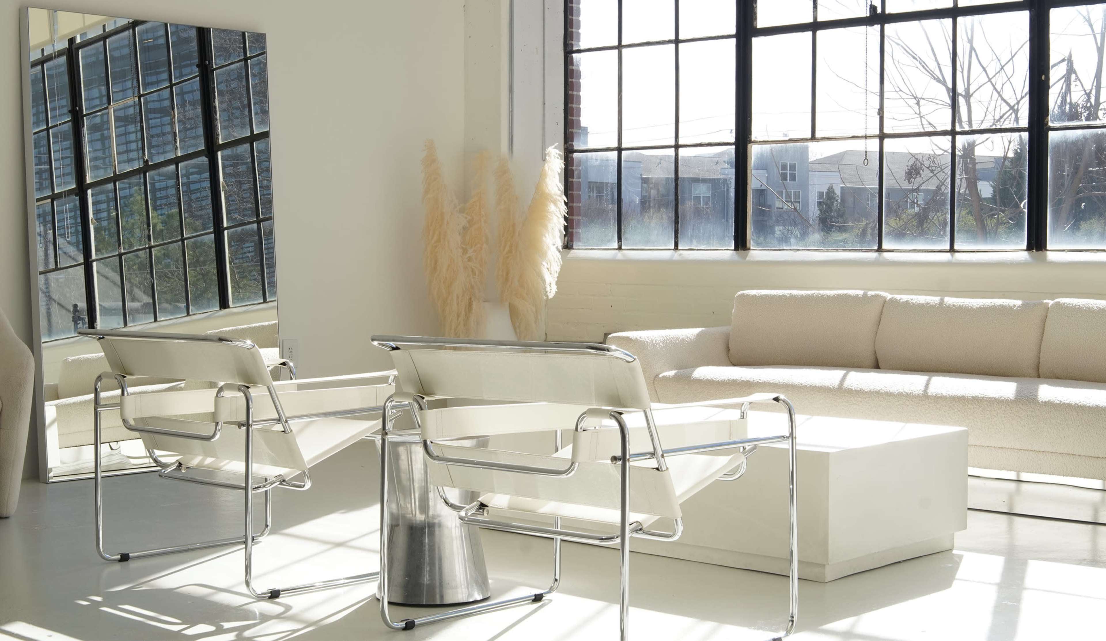 The scene features a bright, minimalist room with large windows, a light-colored sofa, two transparent chairs, and a modern coffee table.