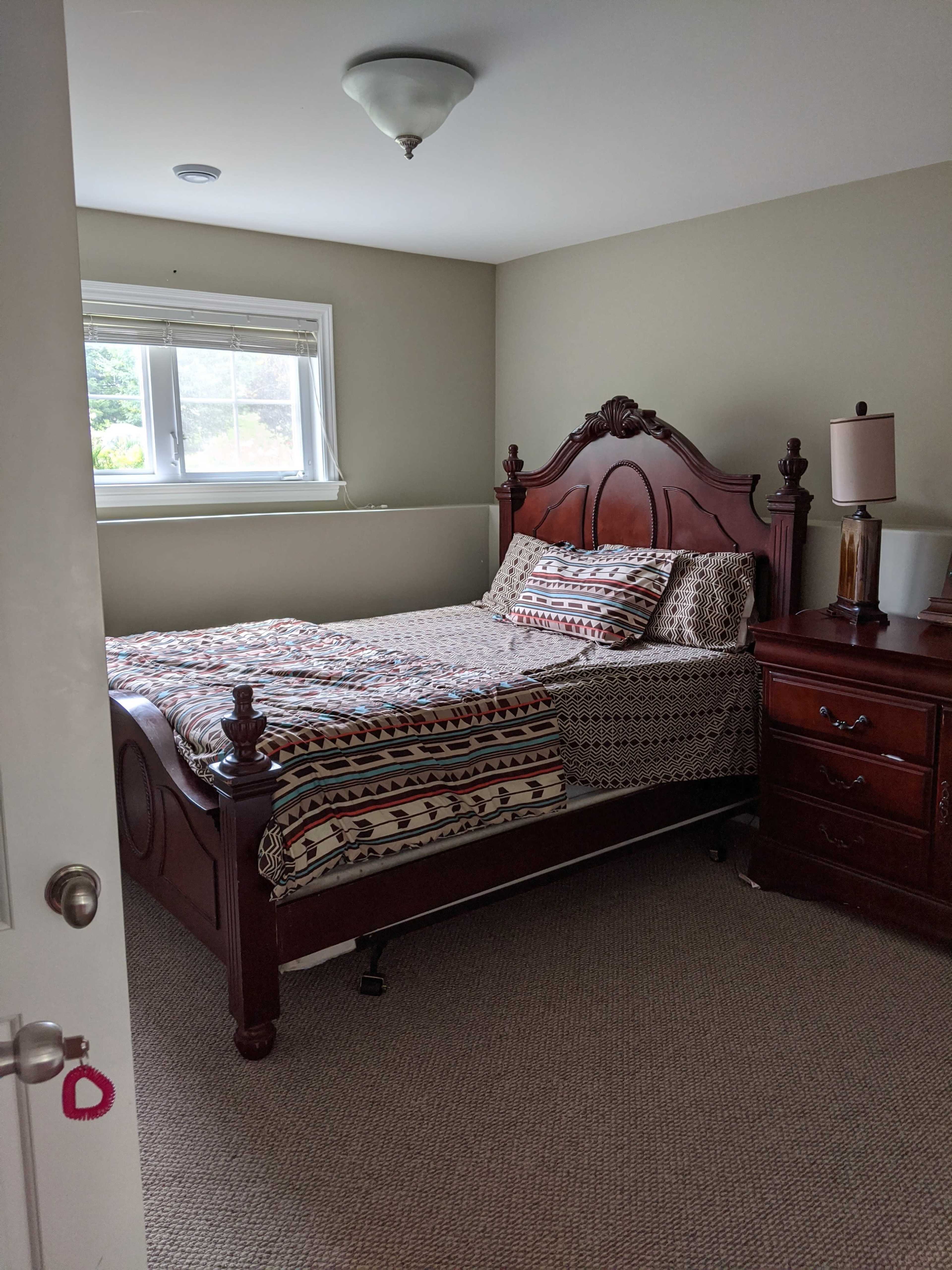 The image shows a neatly arranged bedroom featuring a wooden bed with a patterned comforter and a bedside table with a lamp.