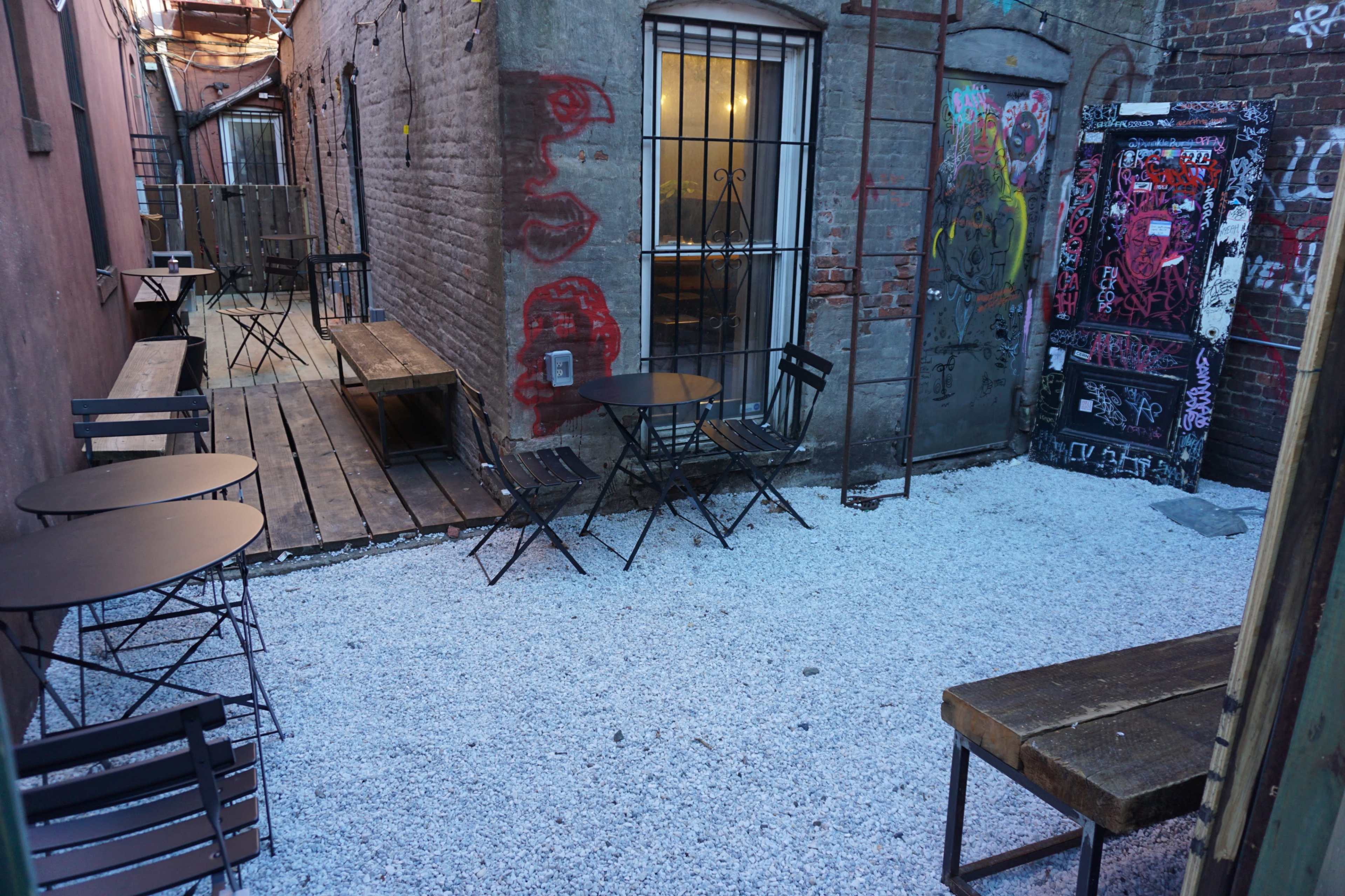 An alleyway with wooden and gravel surfaces, featuring metal tables and chairs surrounded by brick walls adorned with graffiti.