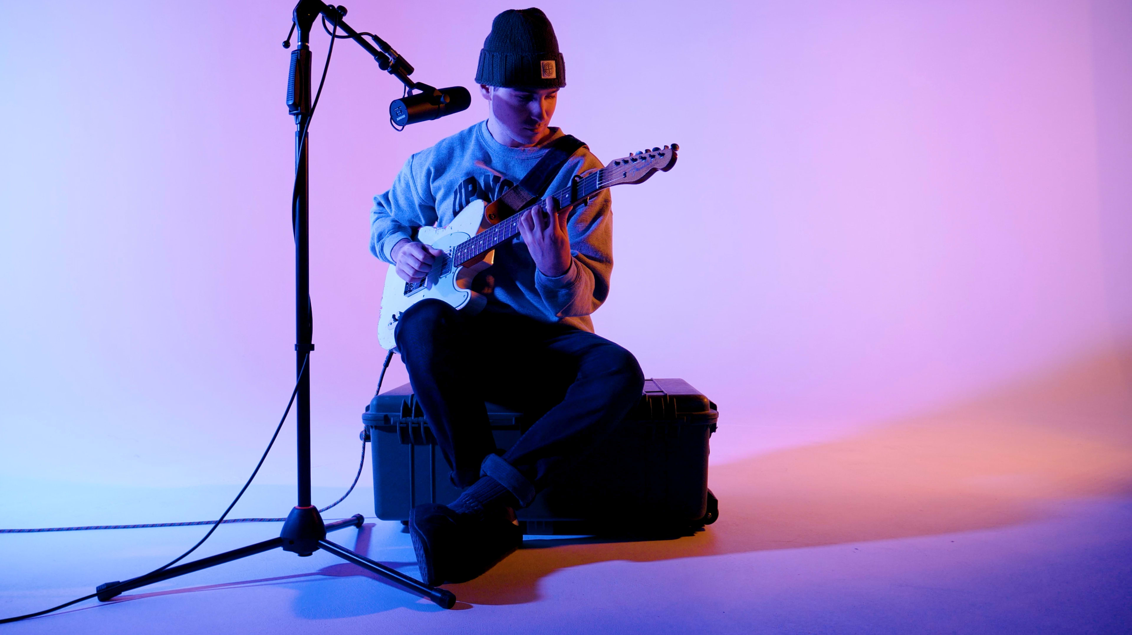 A musician sits on a storage box, playing an electric guitar while facing a microphone on a stand, with a colorful backdrop.