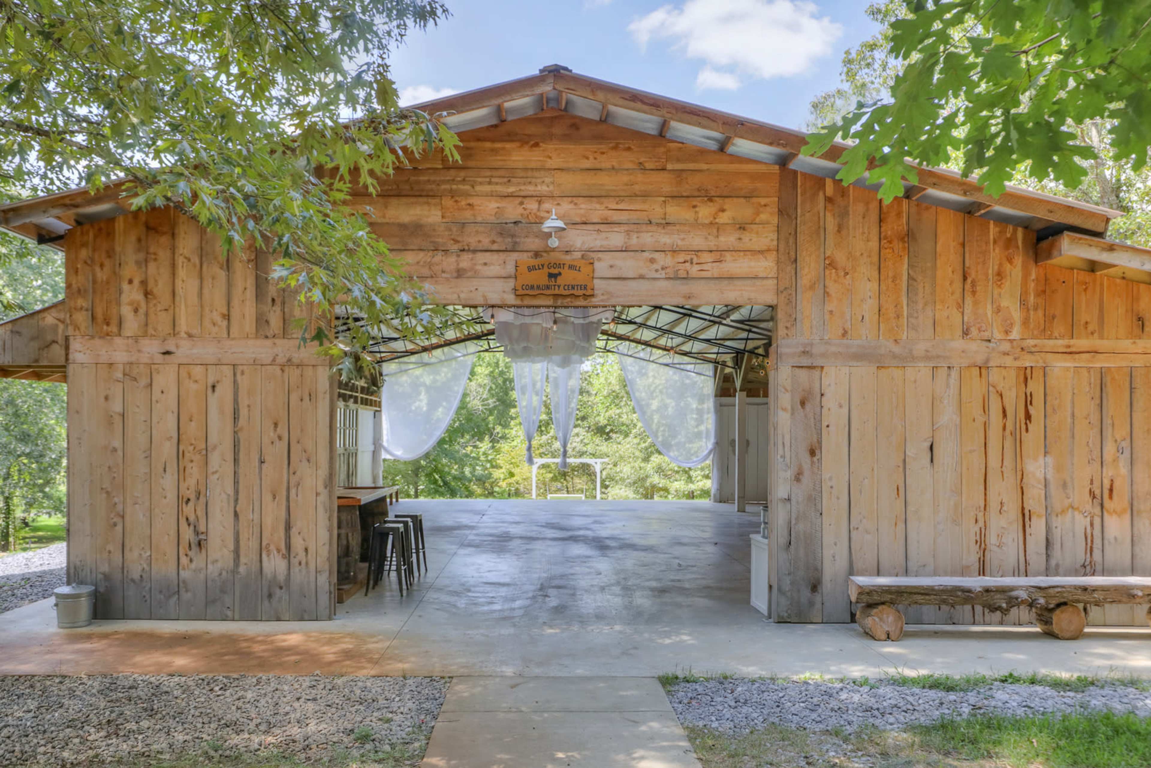 A rustic wooden barn with an open entrance, surrounded by greenery, featuring a concrete floor and scattered seating arrangements inside.