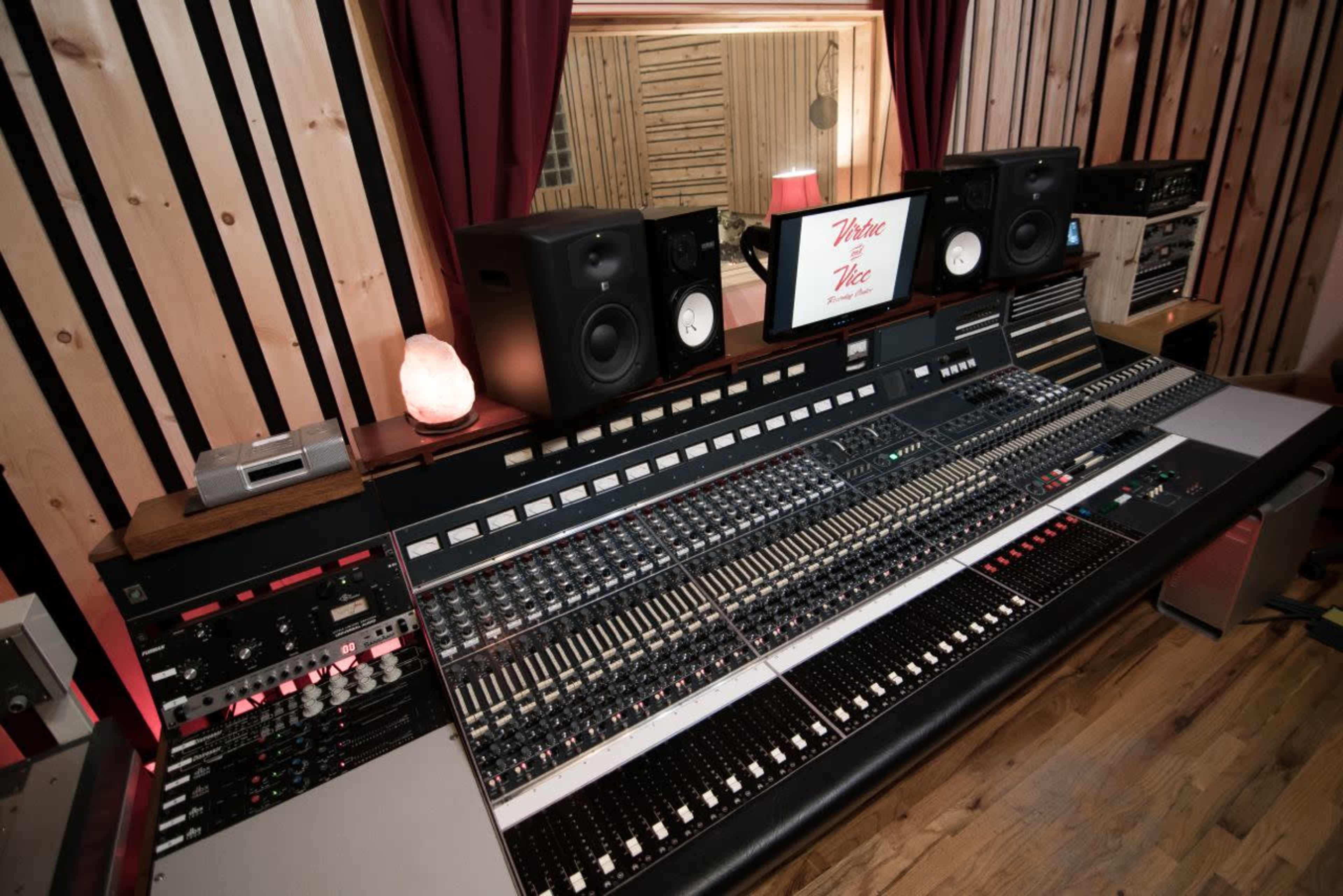 A recording studio control desk with an array of buttons, sliders, and monitors, set against a backdrop of wooden paneling.