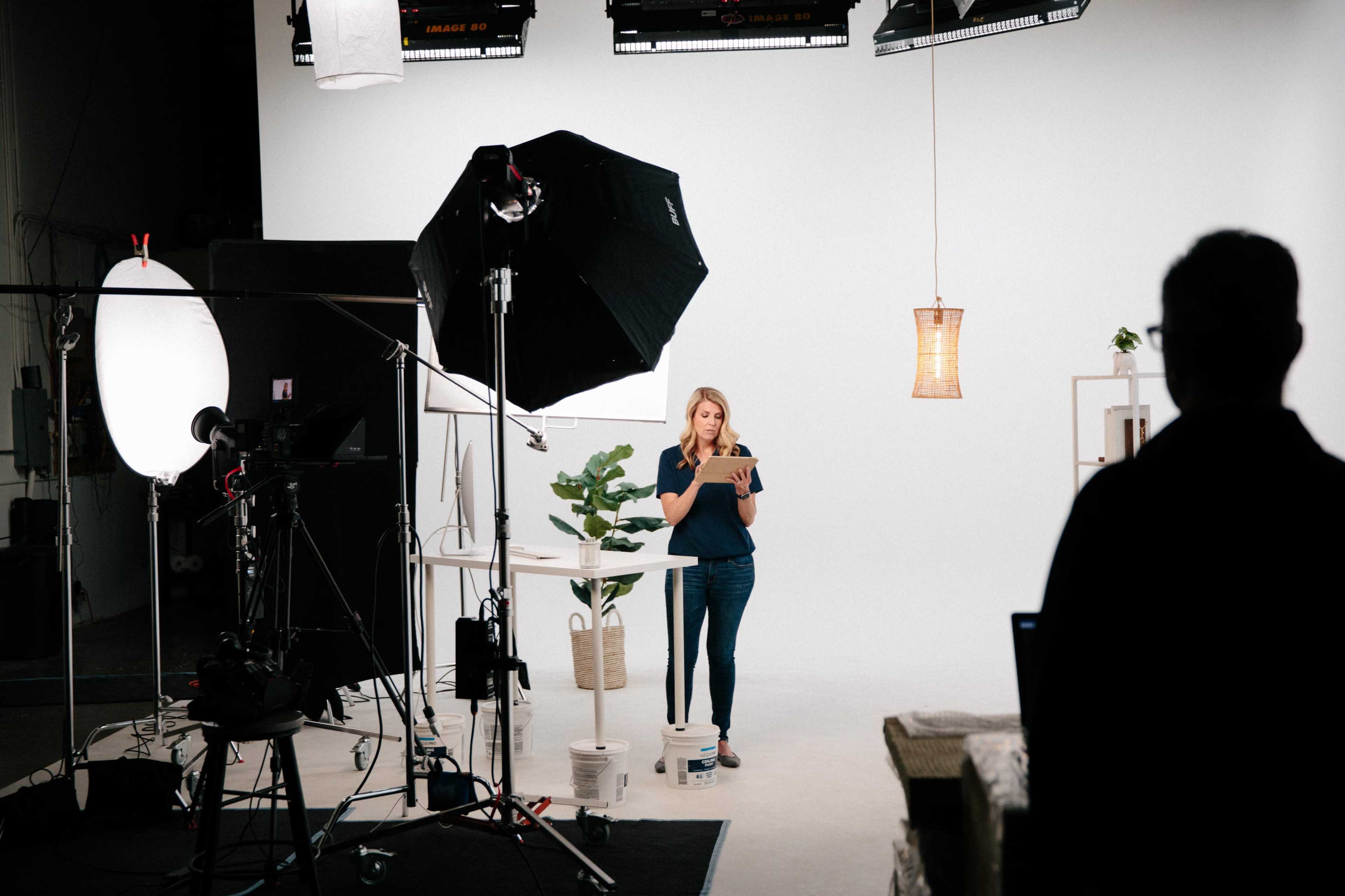 A woman stands on a set with various studio lights and camera equipment, holding a tablet while facing a blank white background.