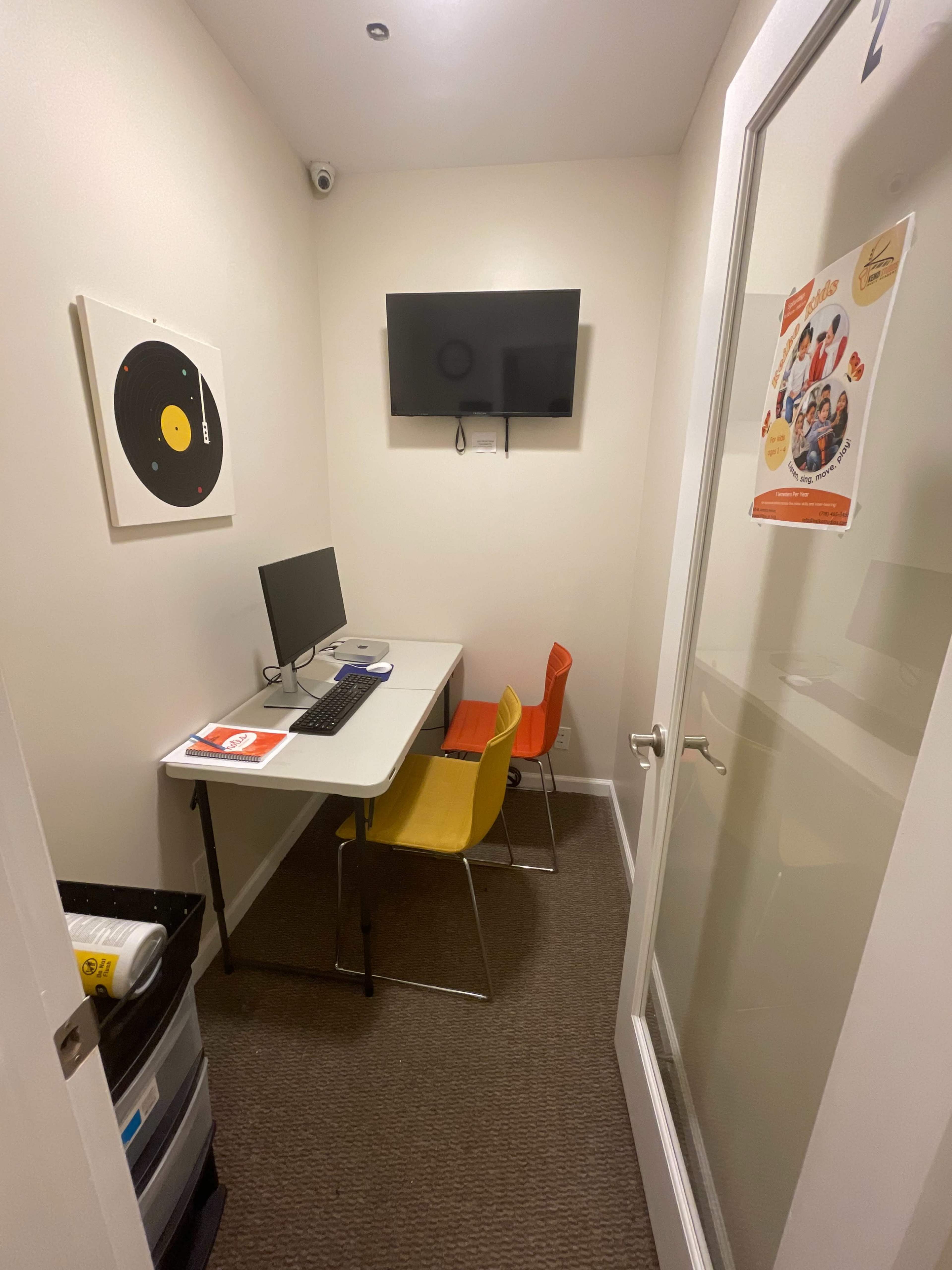 A small, enclosed room features a desk with a computer, two colorful chairs, a television mounted on the wall, and a vinyl record on the wall.