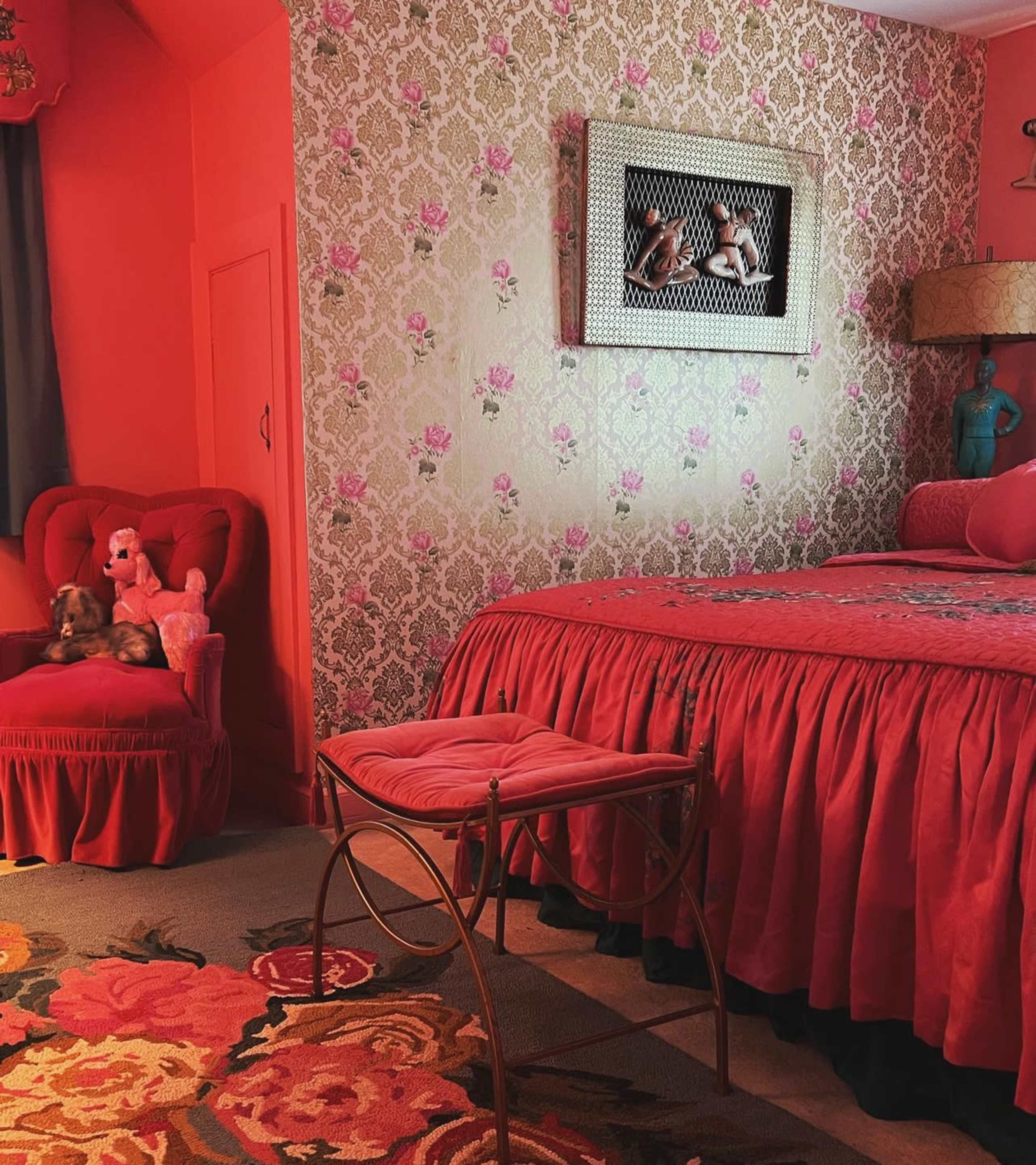 The image shows a vibrant bedroom with a pink floral wallpaper, a bed with a ruffled red bedspread, a small metal stool, and a pink armchair with a stuffed animal.