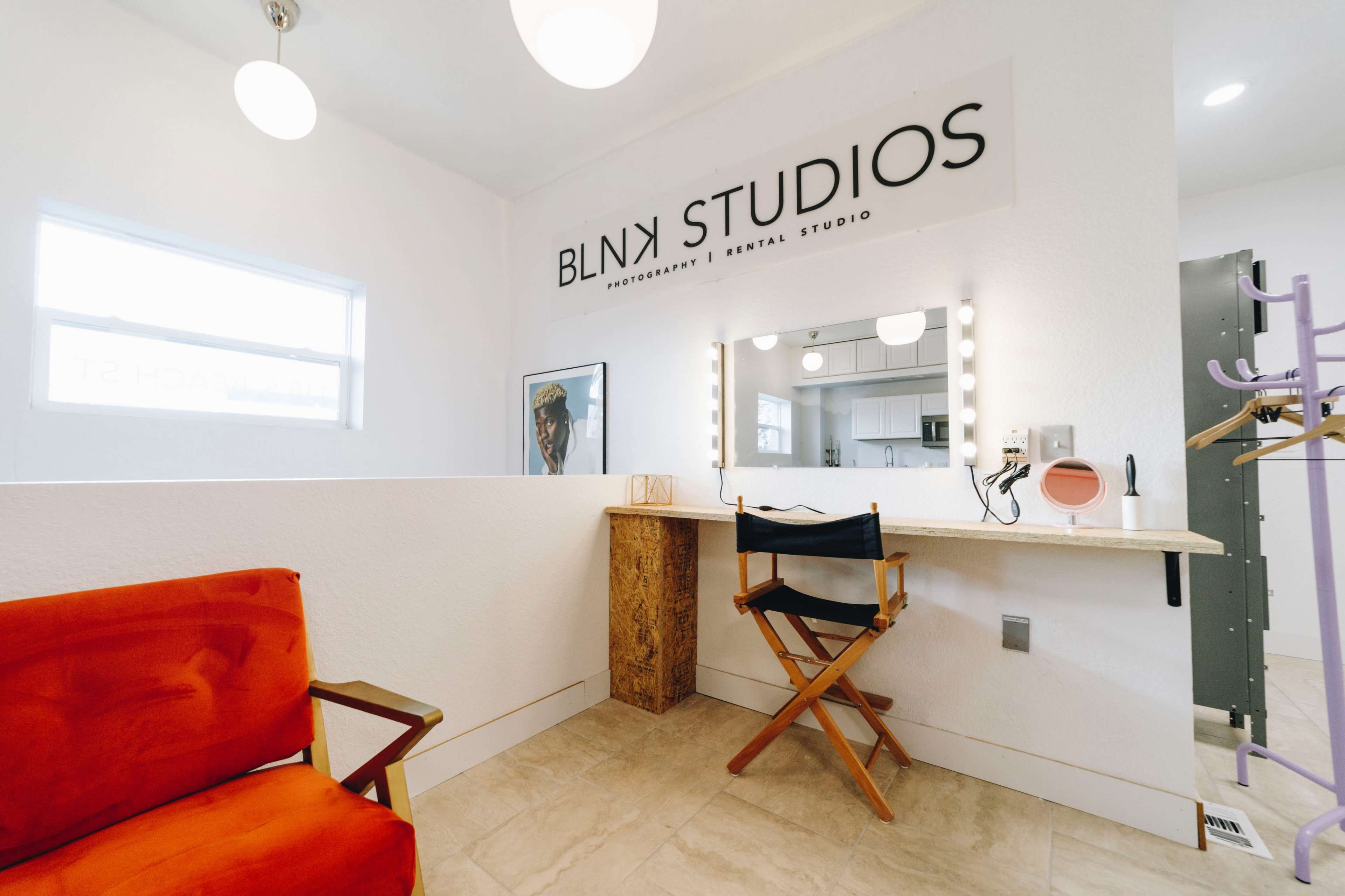 The image shows a well-lit interior of BLNK Studios, featuring a makeup station with bright vanity lights and a red chair, along with a small seating area in the background.