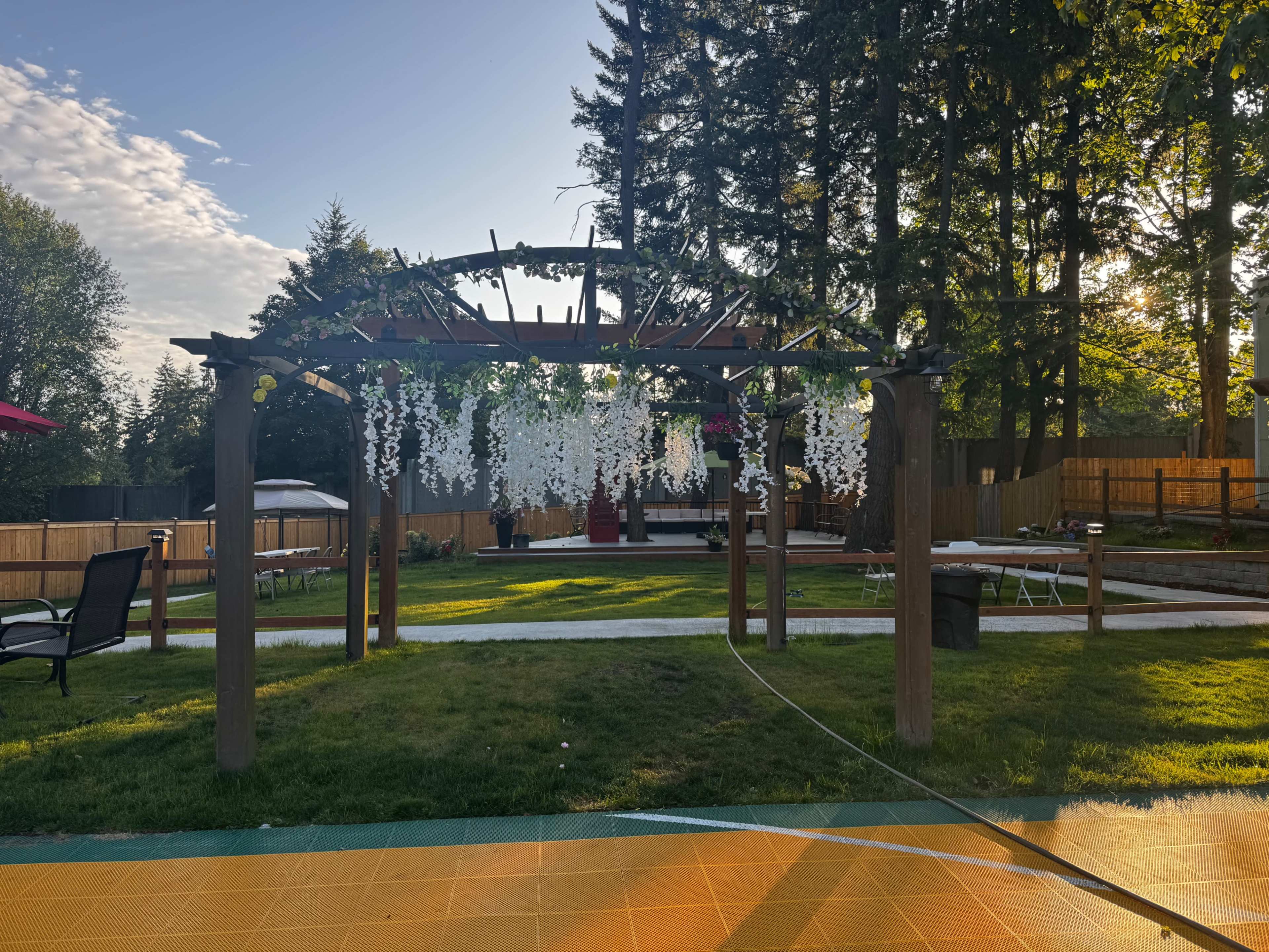 A pergola with hanging flowers is situated in a backyard surrounded by green grass and trees, with a wooden deck and patio in the background.