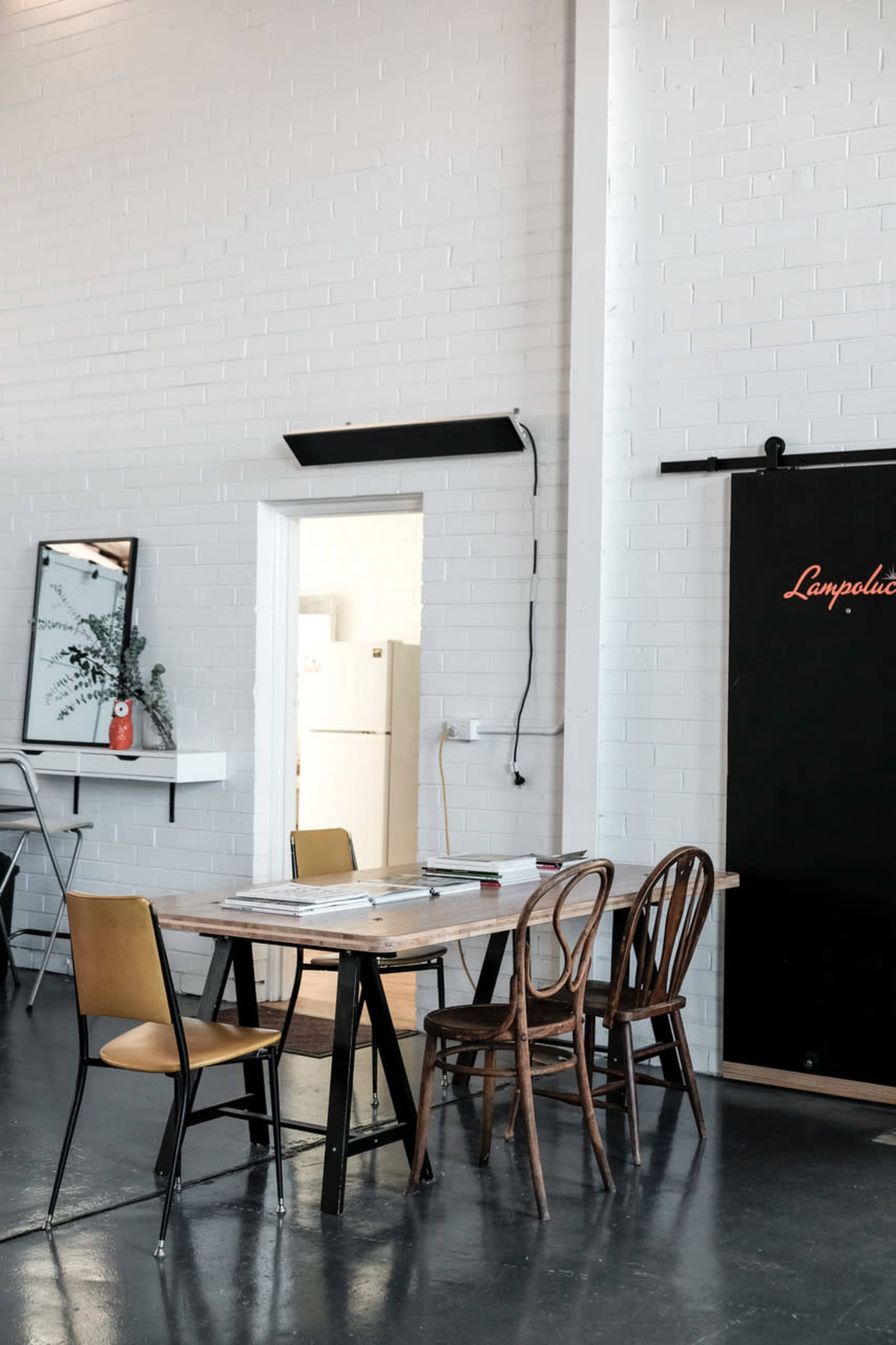 A table with four chairs is set up in a brightly lit room with white brick walls, featuring a blackboard and a doorway leading to another space.